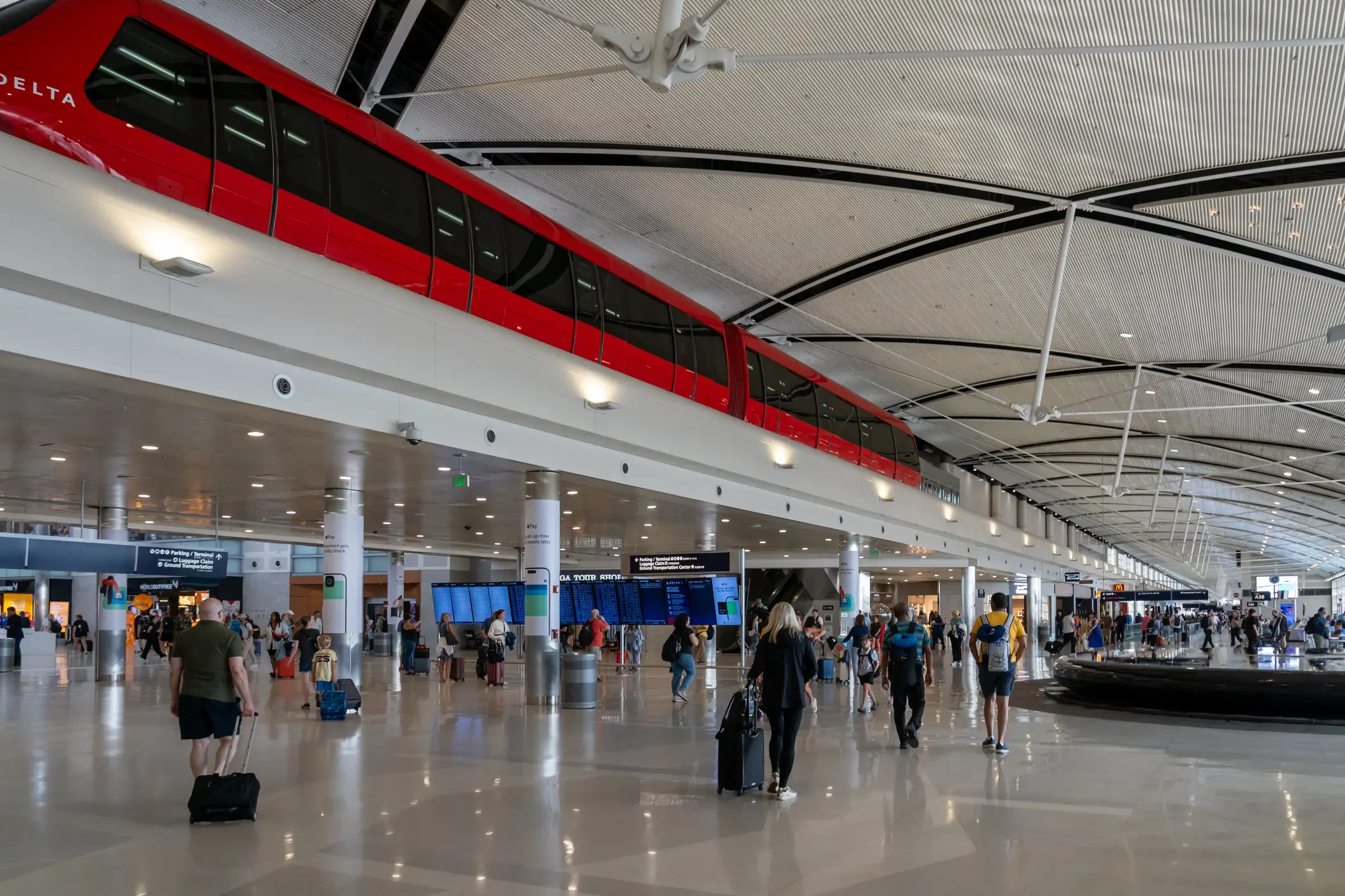 Travelers with luggage move through a bright airport terminal beneath a red DELTA train on an elevated track.