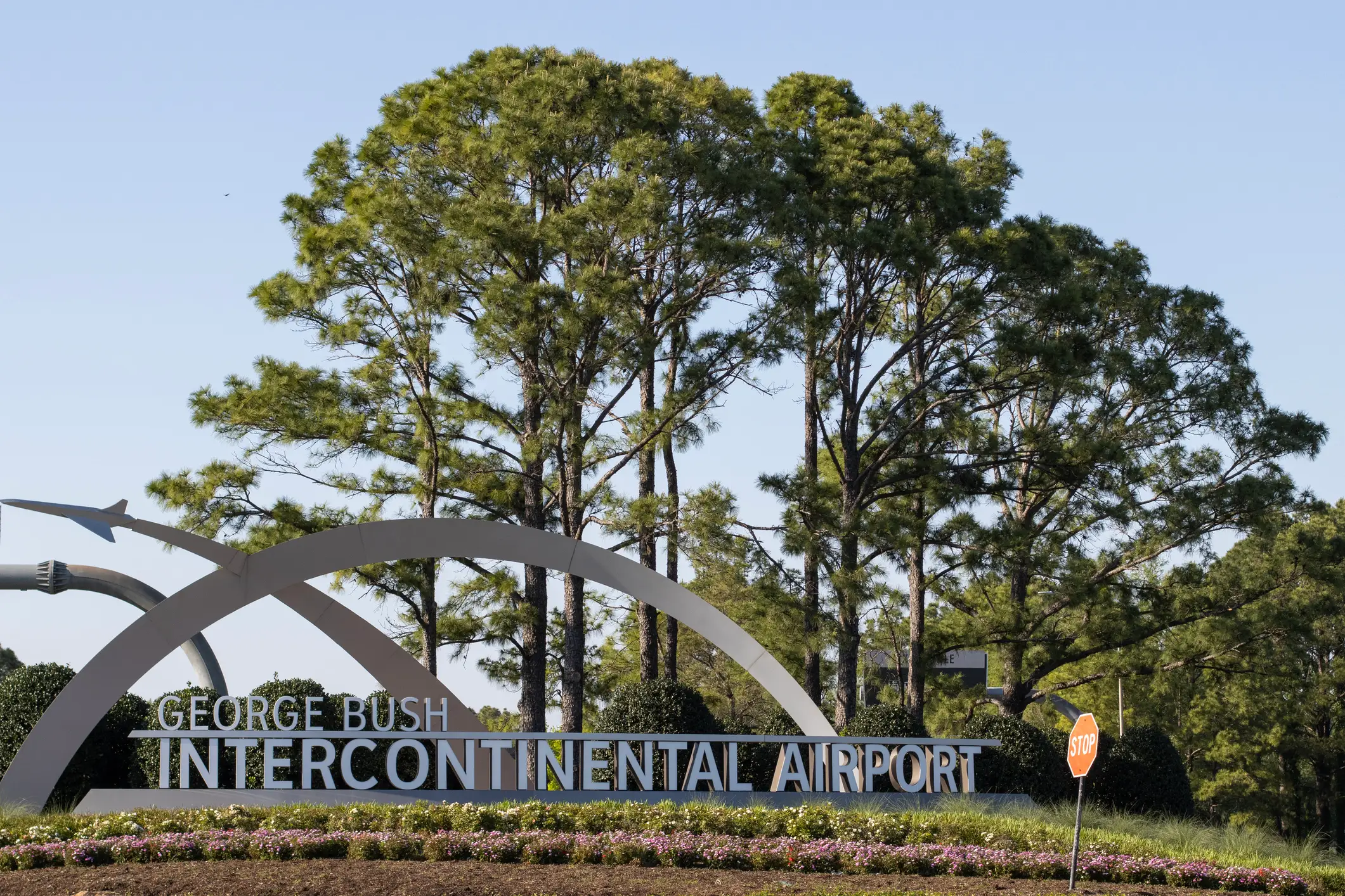 George Bush Intercontinental Airport entrance sign stands beside trees, landscaping, and a stop sign under a clear sky.