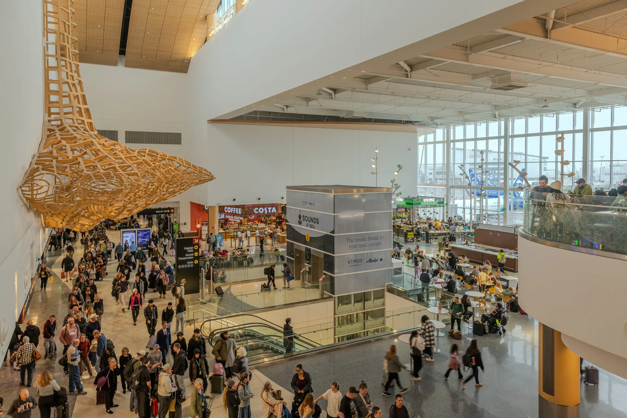 Crowded airport terminal concourse at Seattle-Tacoma International Airport with travelers, a large woven overhead sculpture, cafe seating, escalators, and retail signs.