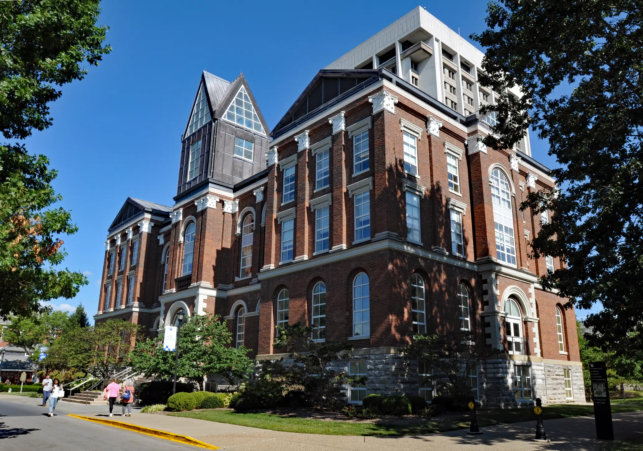 Large red brick institutional building with arched windows stands along a tree-lined campus walkway under a clear blue sky.