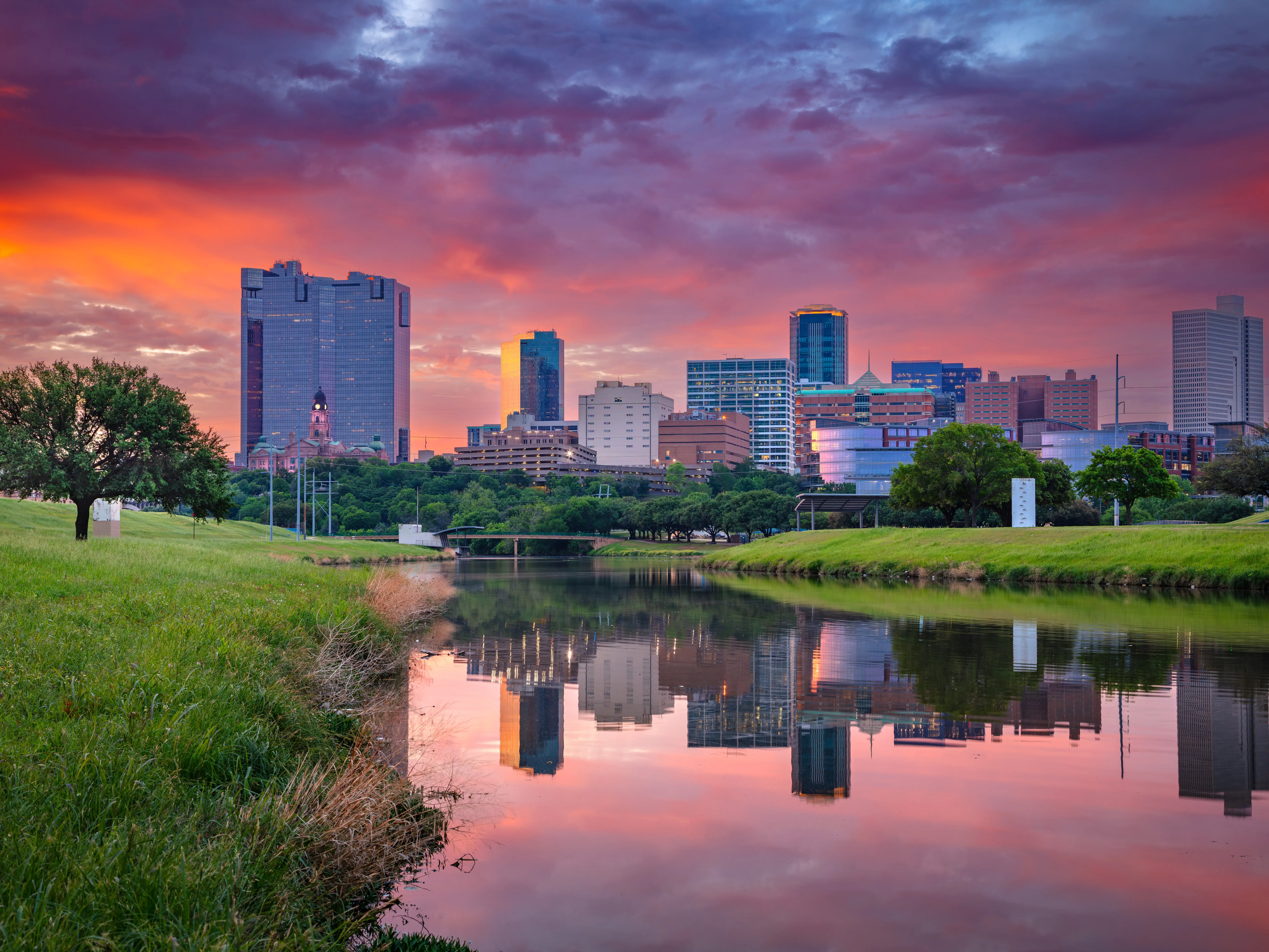 Downtown Fort Worth, Texas, at sunrise. The cityscape features modern skyscrapers reflecting a fiery orange and purple sky. The Trinity River in the foreground mirrors the skyline and vibrant clouds. Lush green grass and a few trees line the riverbanks, adding a natural element to the urban scene. Prominent buildings rise against the horizon, showcasing the architecture of this Texan city.