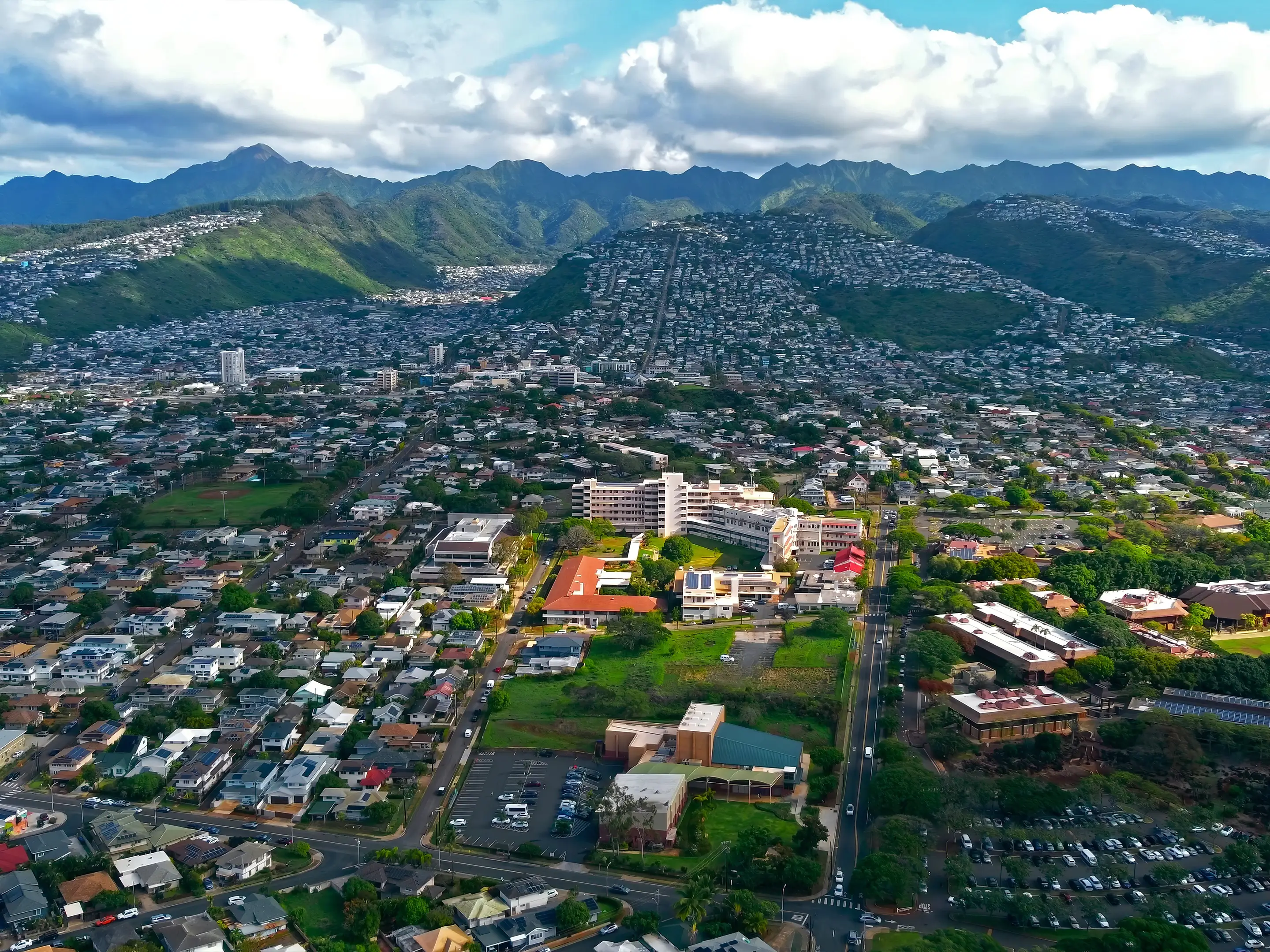 Aerial View of Honolulu Neighborhoods and Mountains. Wide aerial view of residential neighborhoods in Honolulu, Hawaii, with lush green mountains and scattered clouds in the background