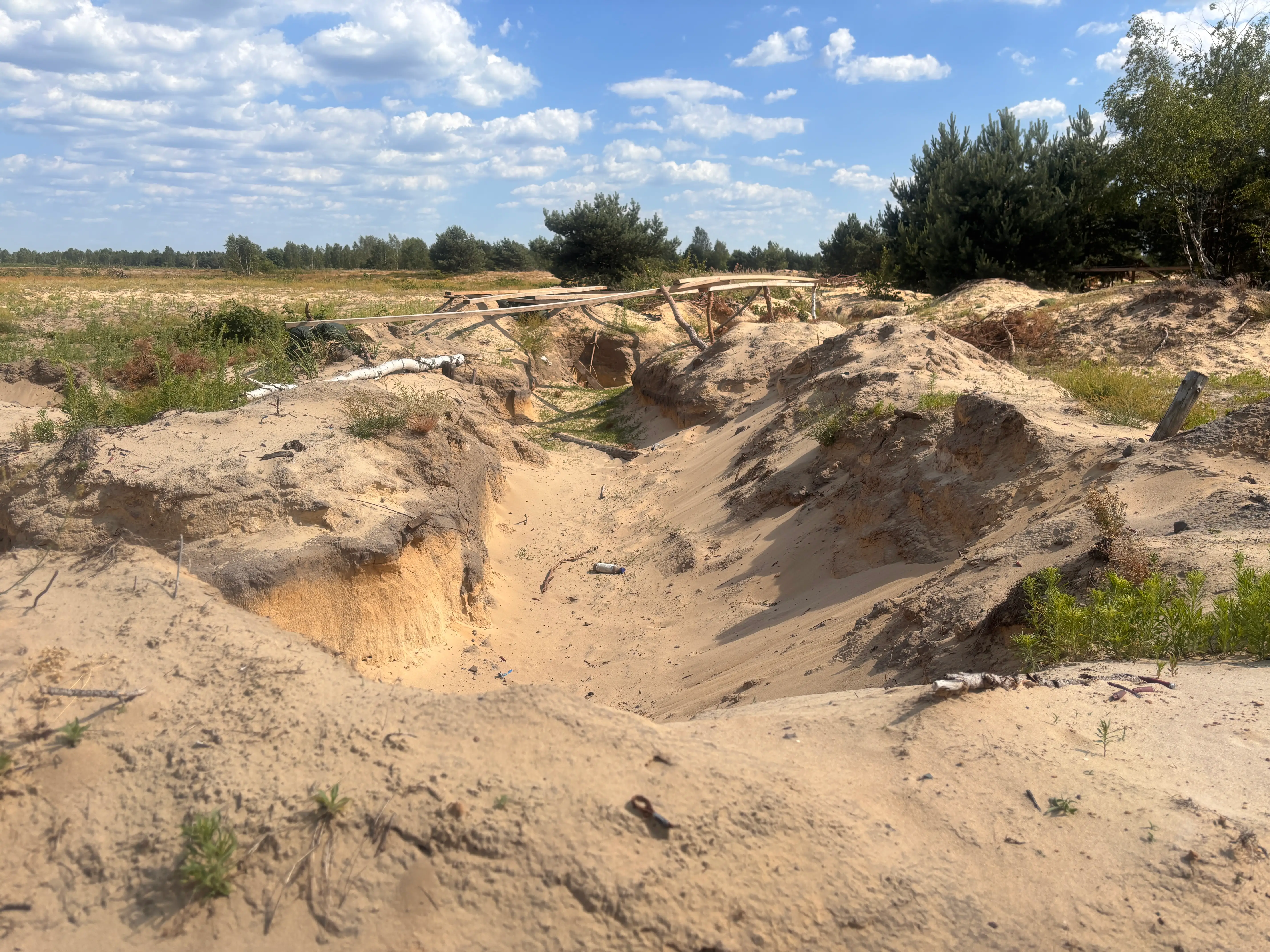 Sandy eroded trench with makeshift wooden plank crossings in a sparse rural landscape under a cloudy blue sky.