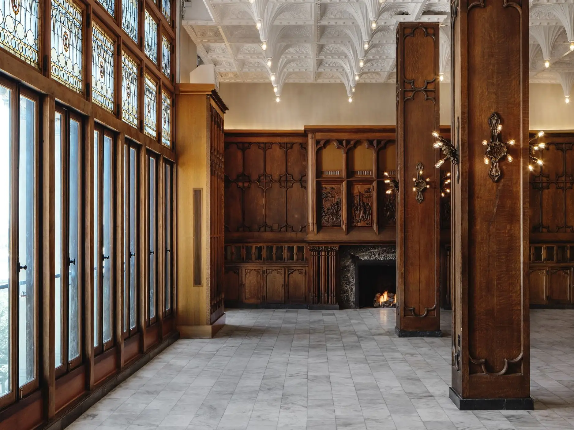 Ornate historic interior with stained-glass windows, carved wood paneling, marble floor, decorative ceiling, and lit fireplace.
