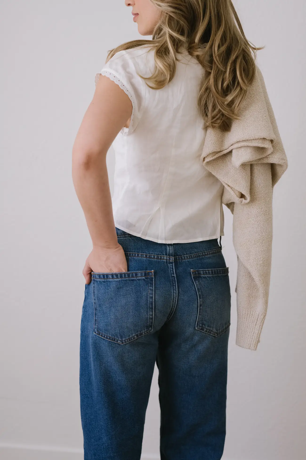 Person with long wavy hair wearing a white blouse and blue jeans, holding a beige sweater over one shoulder.