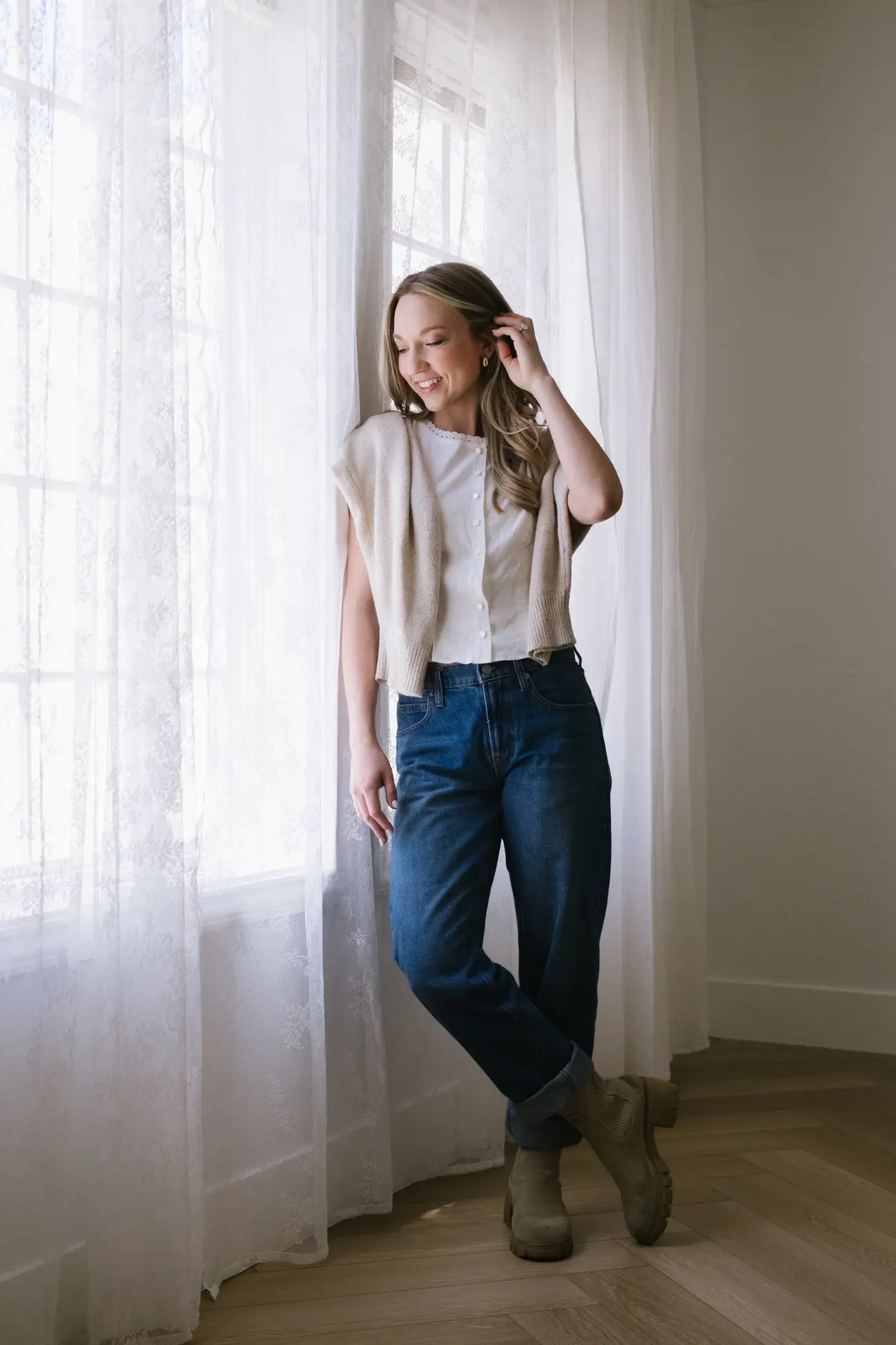 A smiling person stands beside sheer curtained windows in a bright room wearing jeans and layered neutral clothing.