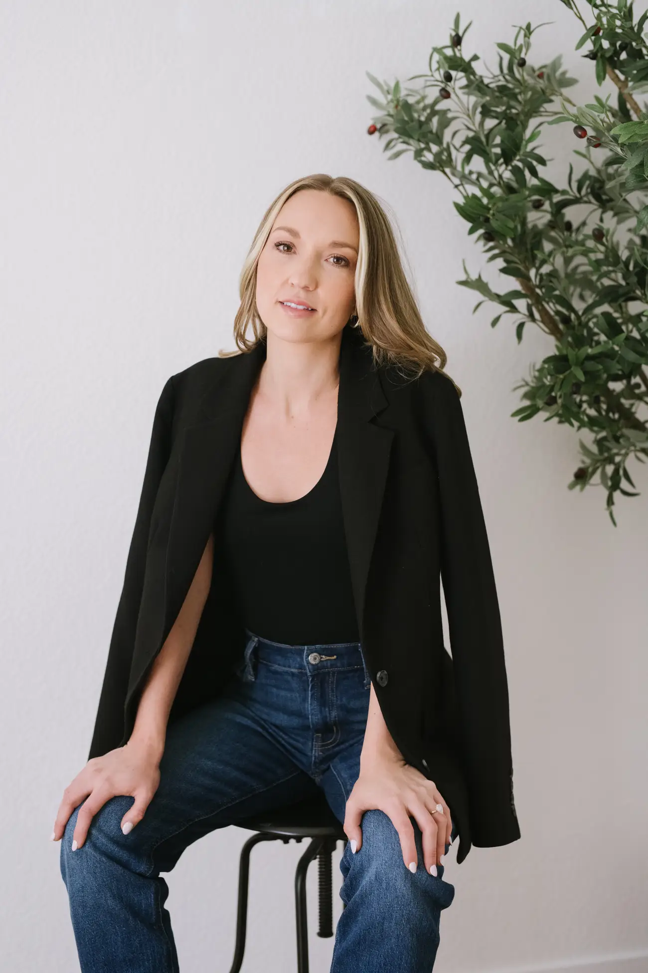 Seated person in a black blazer and jeans poses beside a leafy potted plant against a plain wall.
