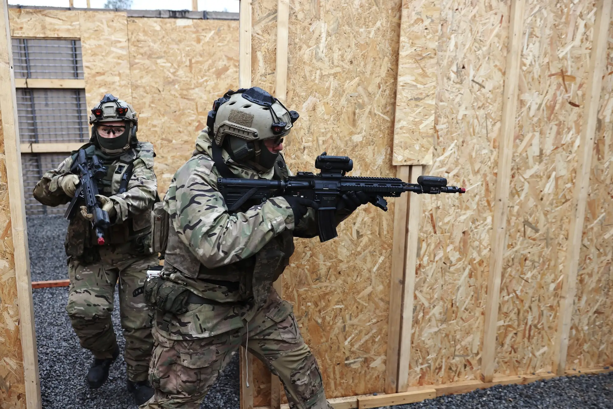 Camouflaged tactical personnel with rifles move through a plywood-walled training structure.