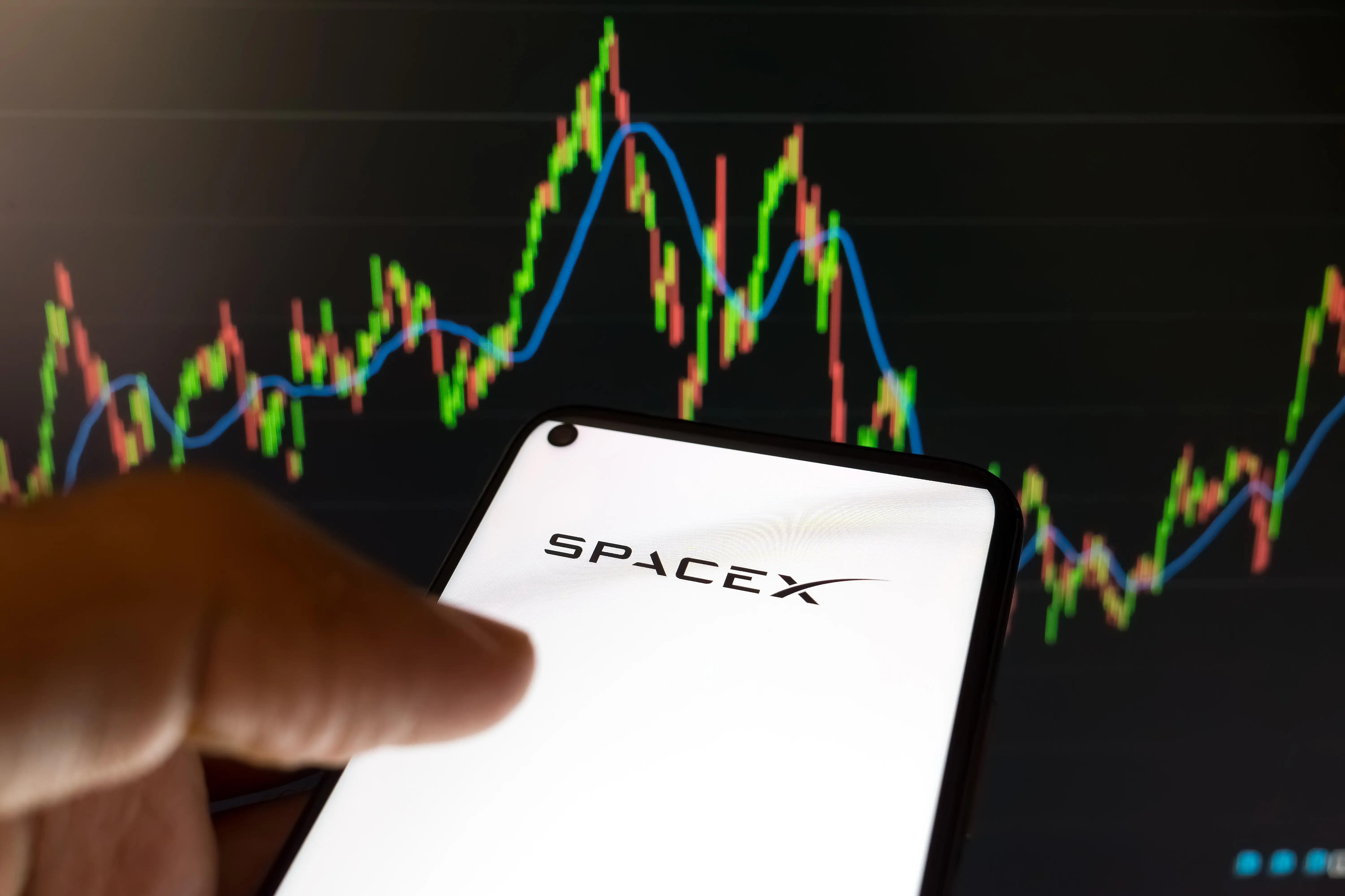 A smartphone displaying the SPACEX logo is held in front of a colorful financial chart on a dark screen.