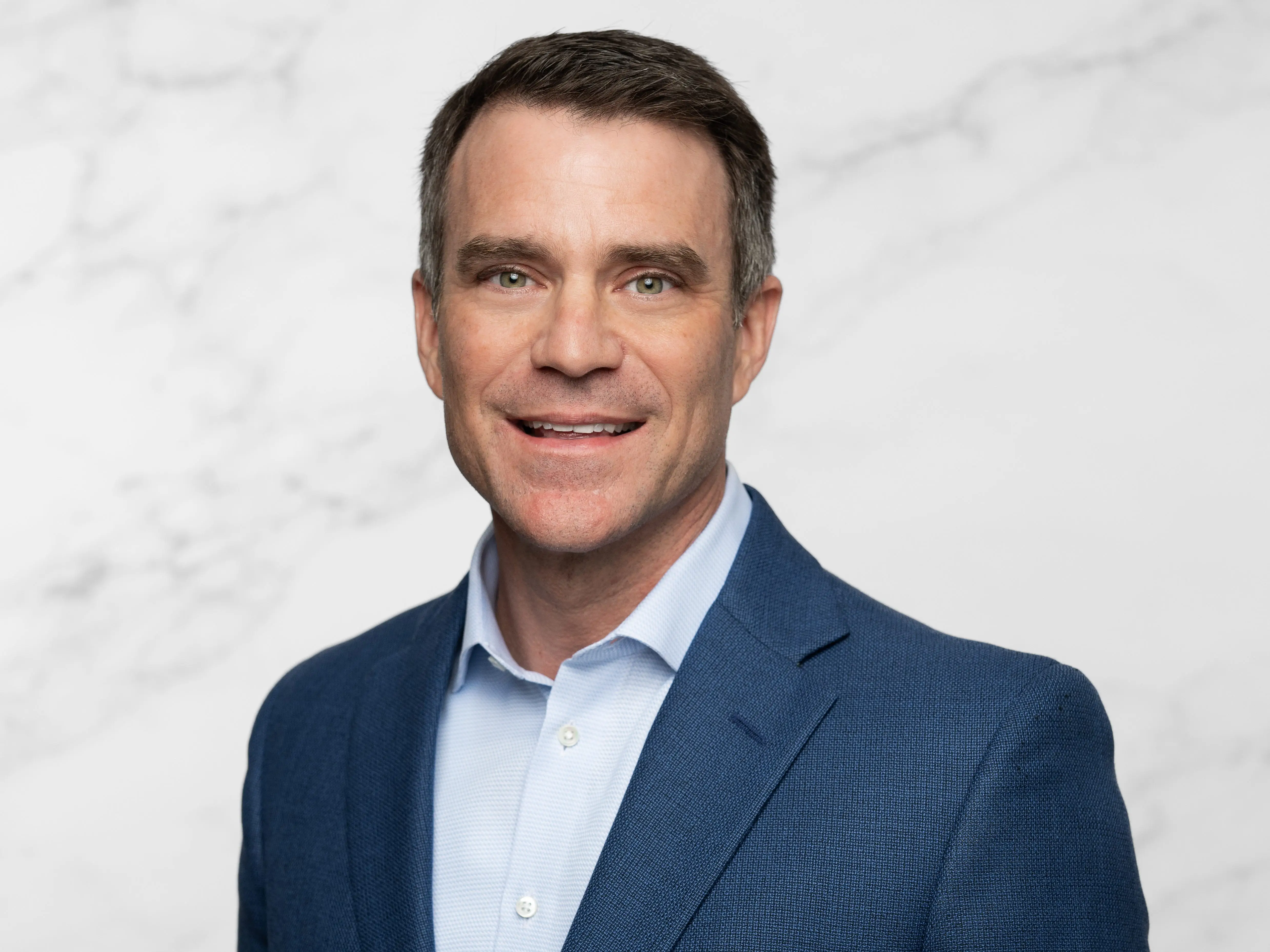 Smiling person in a blue blazer and light blue shirt poses against a pale marble-patterned background.