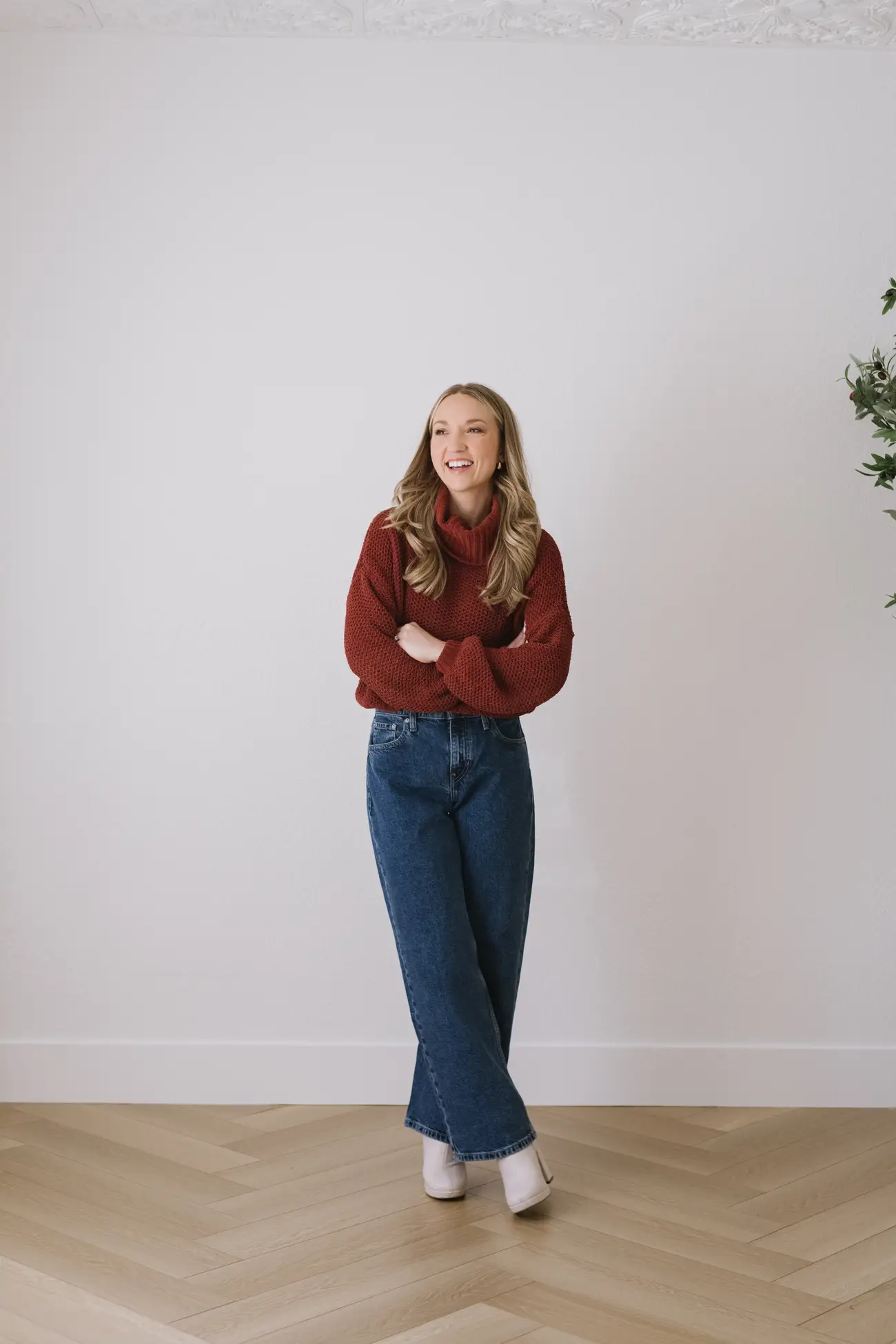 Smiling person in a red turtleneck sweater and jeans stands against a plain white wall.