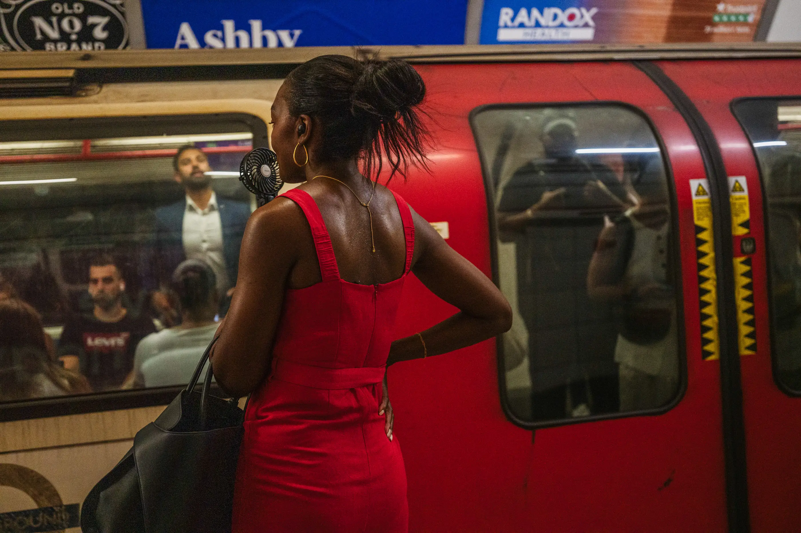 Person in a red dress stands on an Underground platform beside a red train with passengers reflected in the windows.