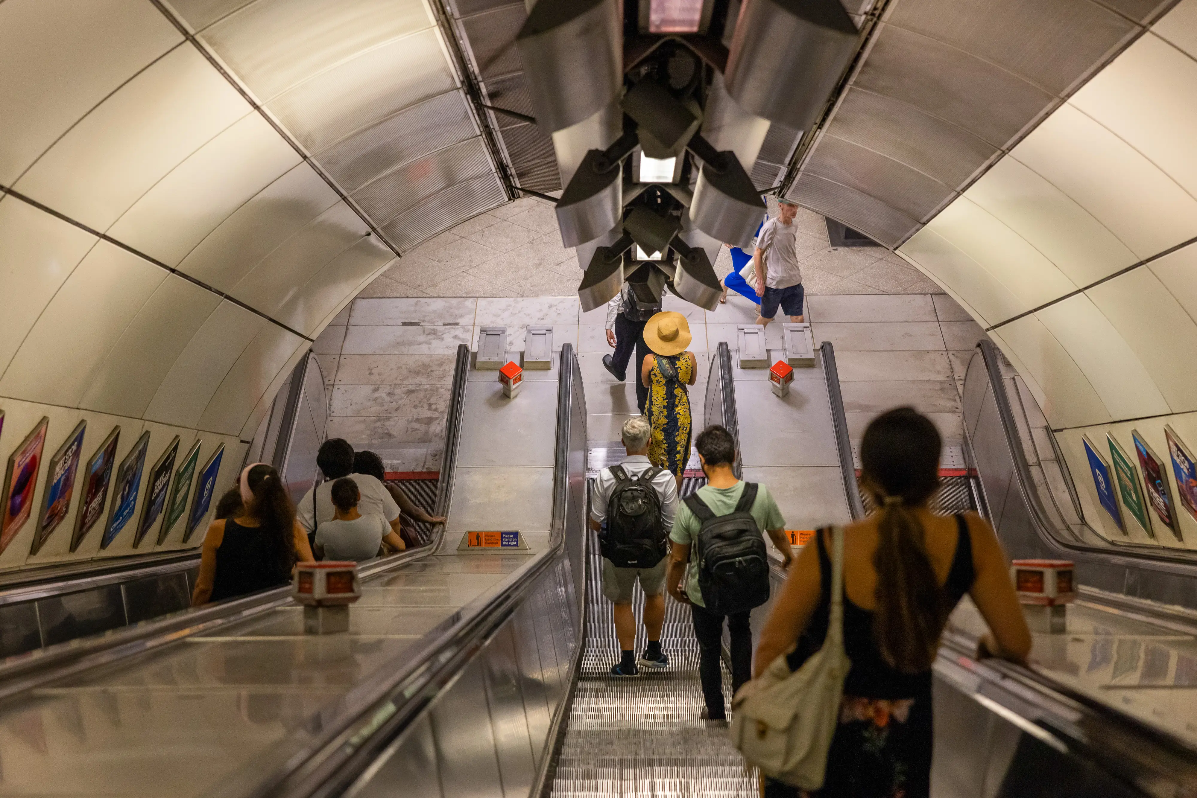 Commuters ride long escalators through a curved underground transit station tunnel with metallic walls.