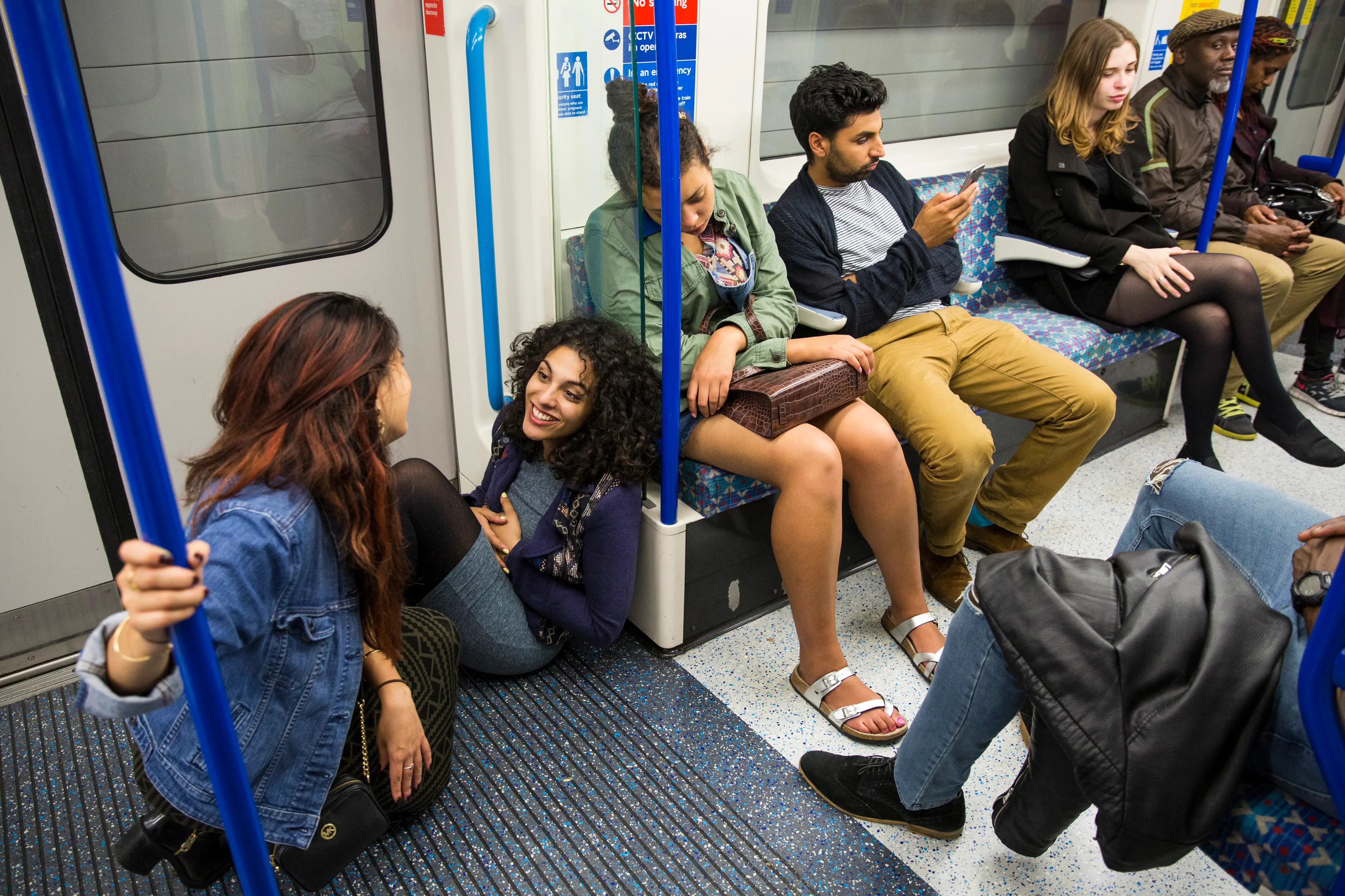 Passengers sit and stand inside a crowded metro train carriage with blue poles and patterned seats.