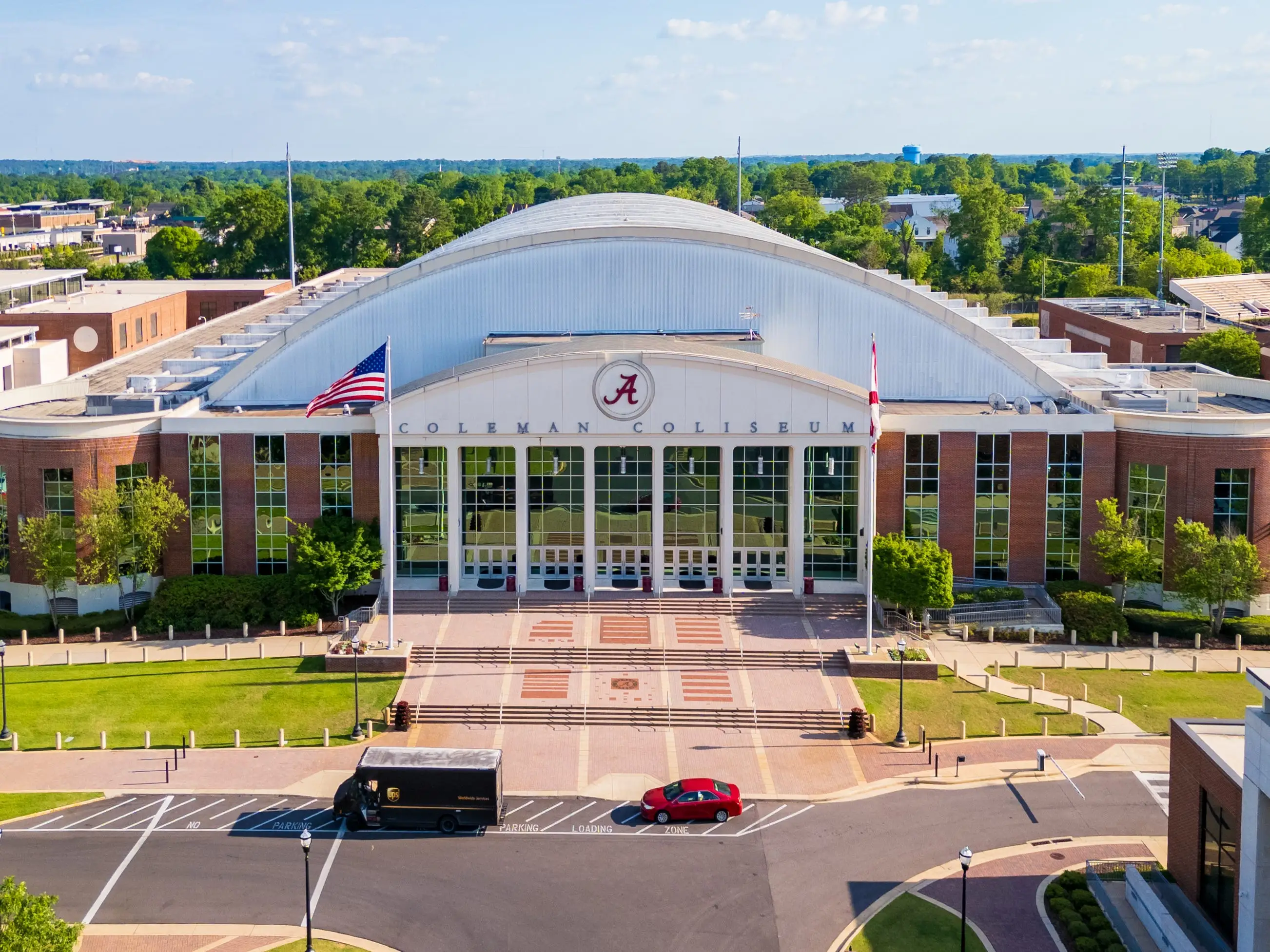 Aerial view of Coleman Coliseum entrance with Alabama logo, flags, plaza, and parked vehicles.