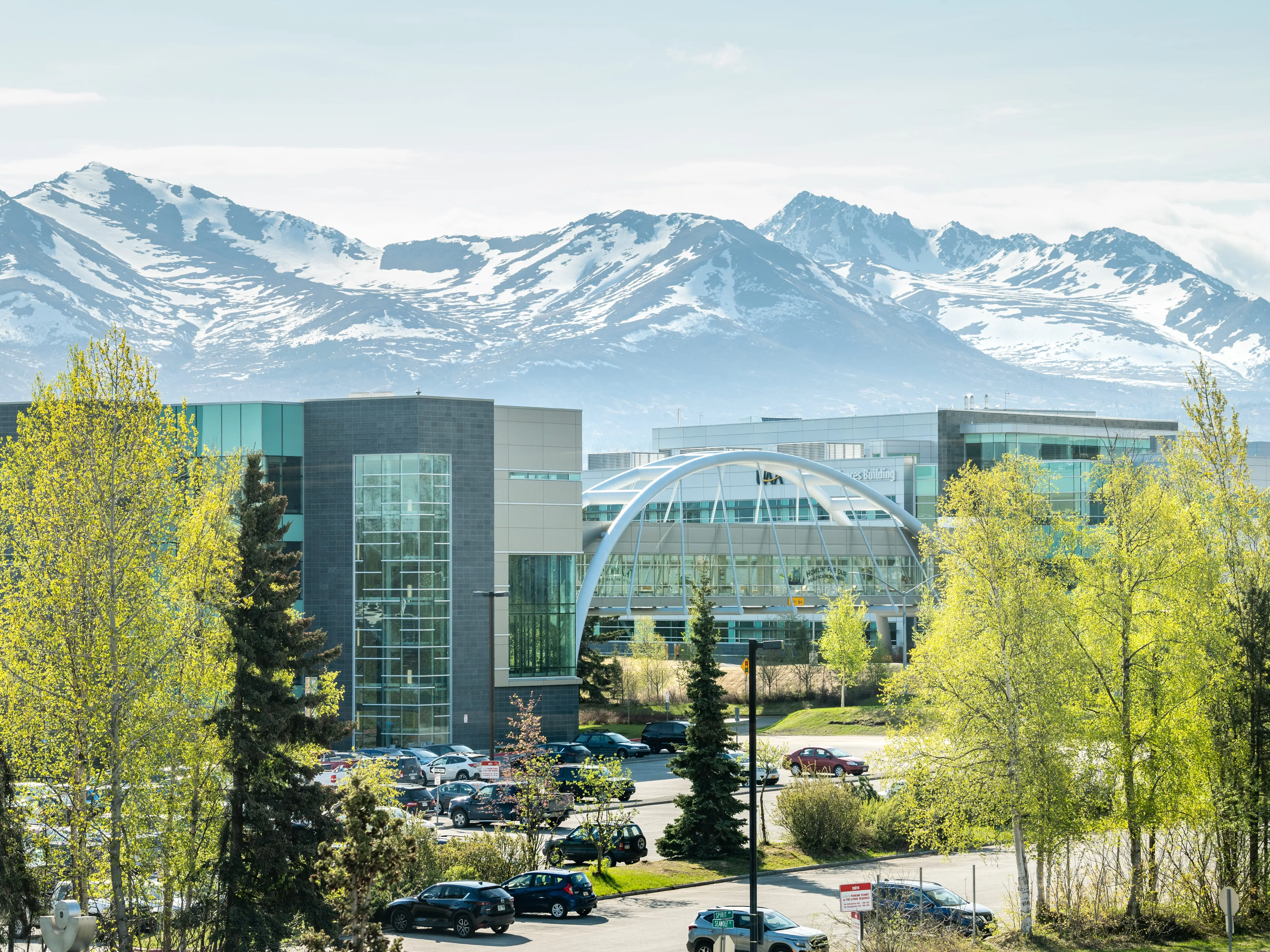 Modern glass buildings and parked cars sit among spring trees with snow-covered mountains in the background.
