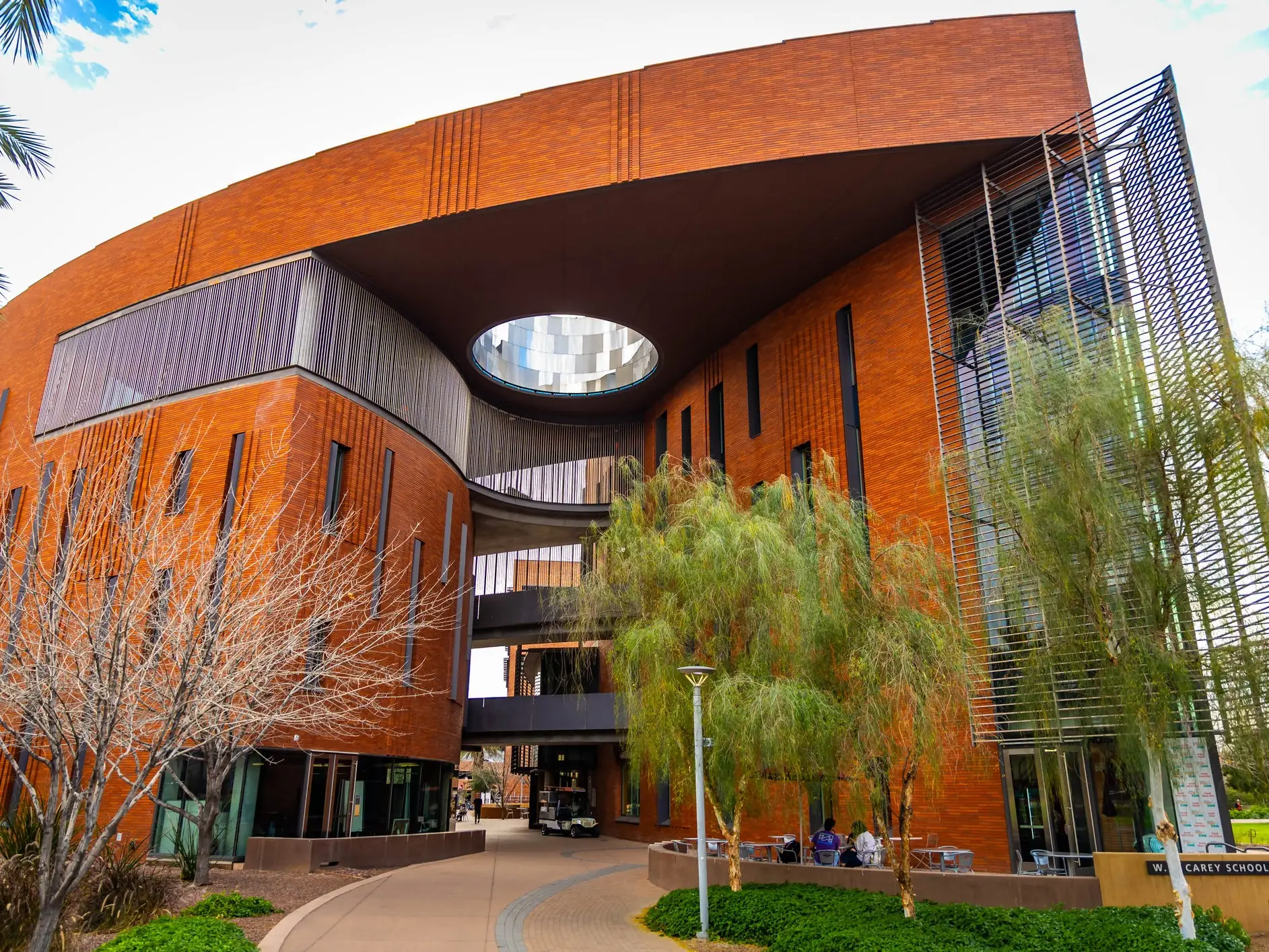 Curved brick W. P. Carey School of Business building with metal screens, courtyard trees, and walkway at Arizona State University.
