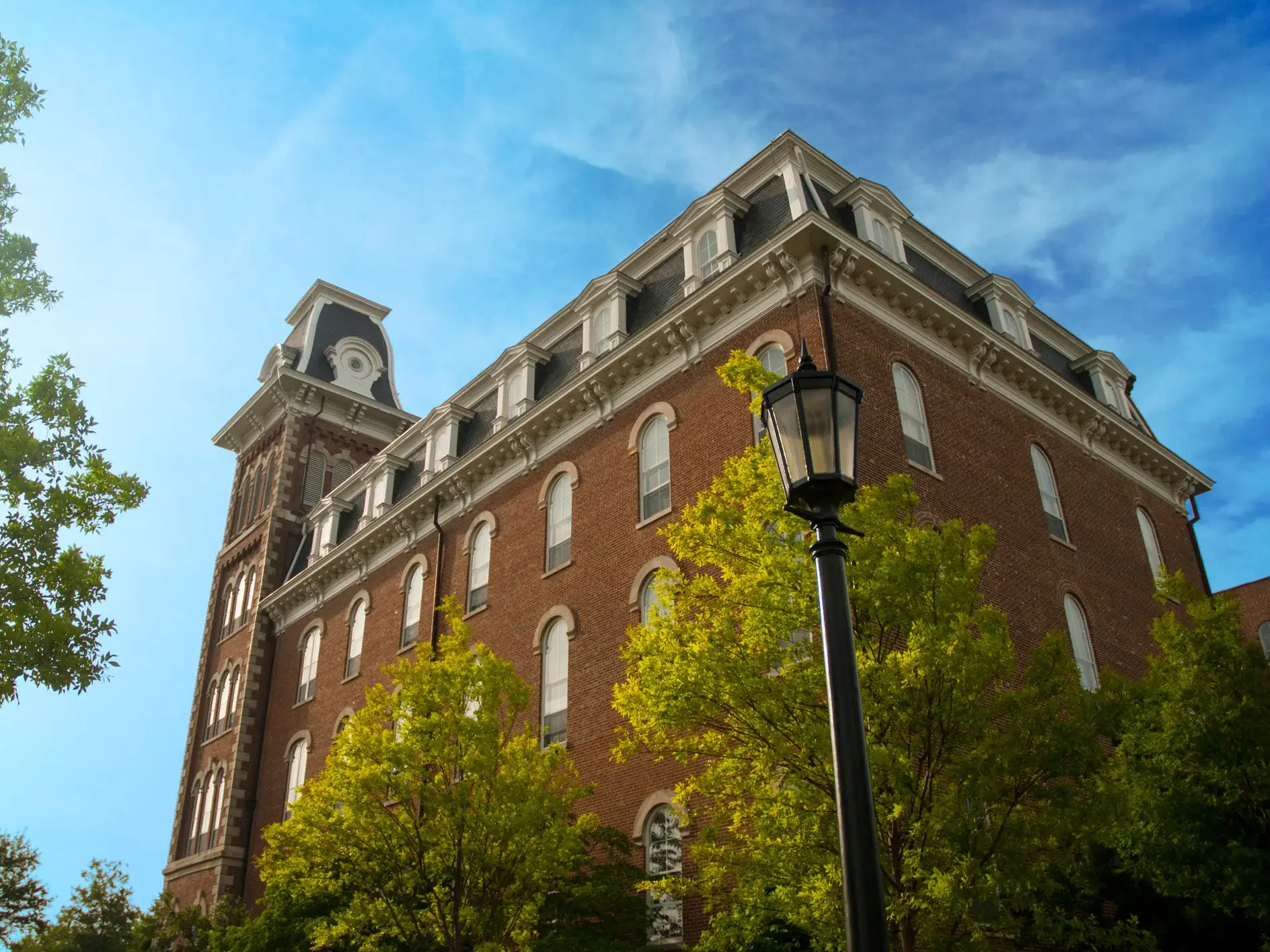 Red brick historic building, Old Main, a landmark and symbol of the University of Arkansas Campus in Arkansas, with leafy trees and a black streetlamp under a blue sky.