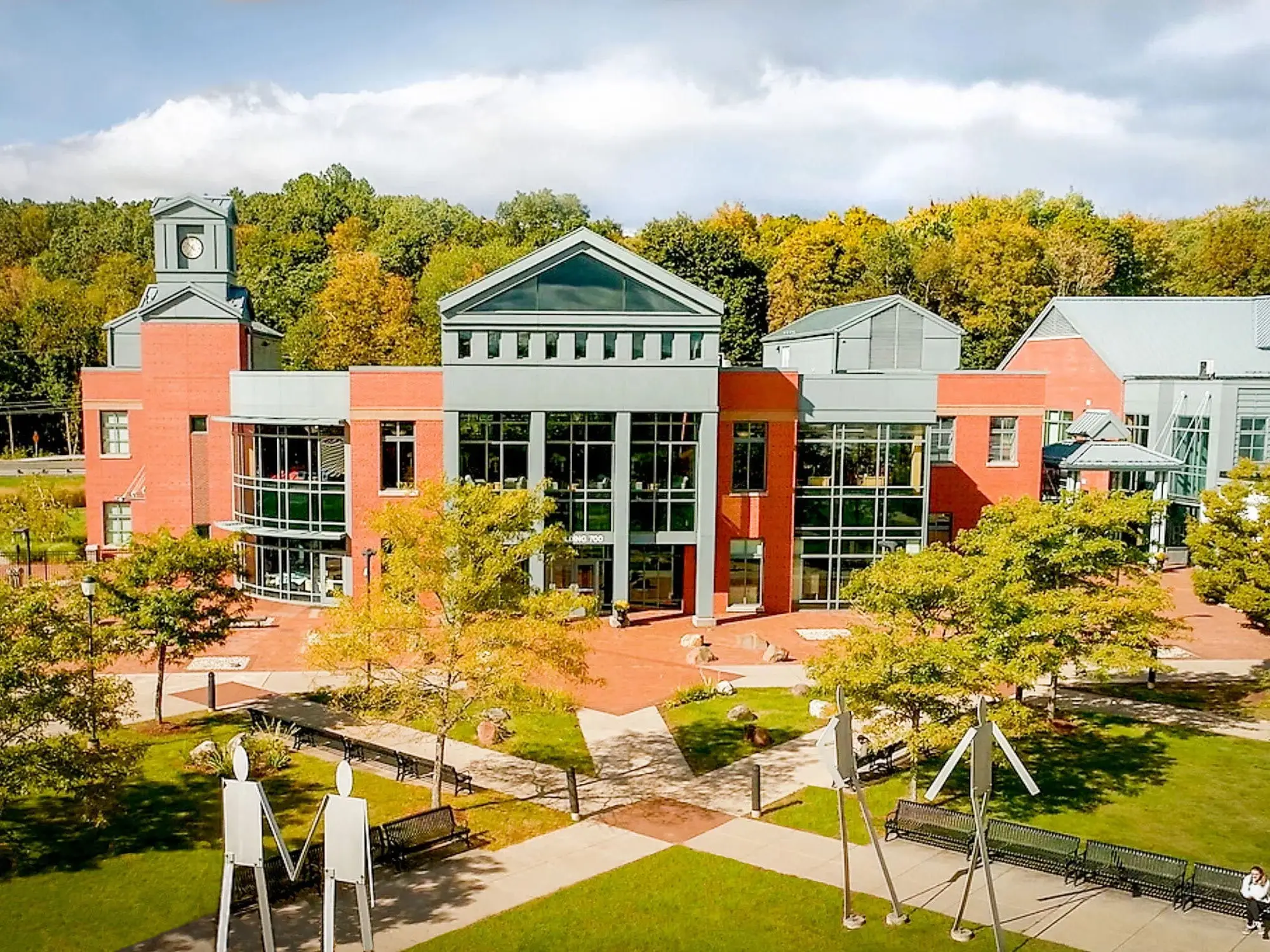 Red brick campus building with glass atrium, clock tower, walkways, benches, and autumn trees under cloudy sky.