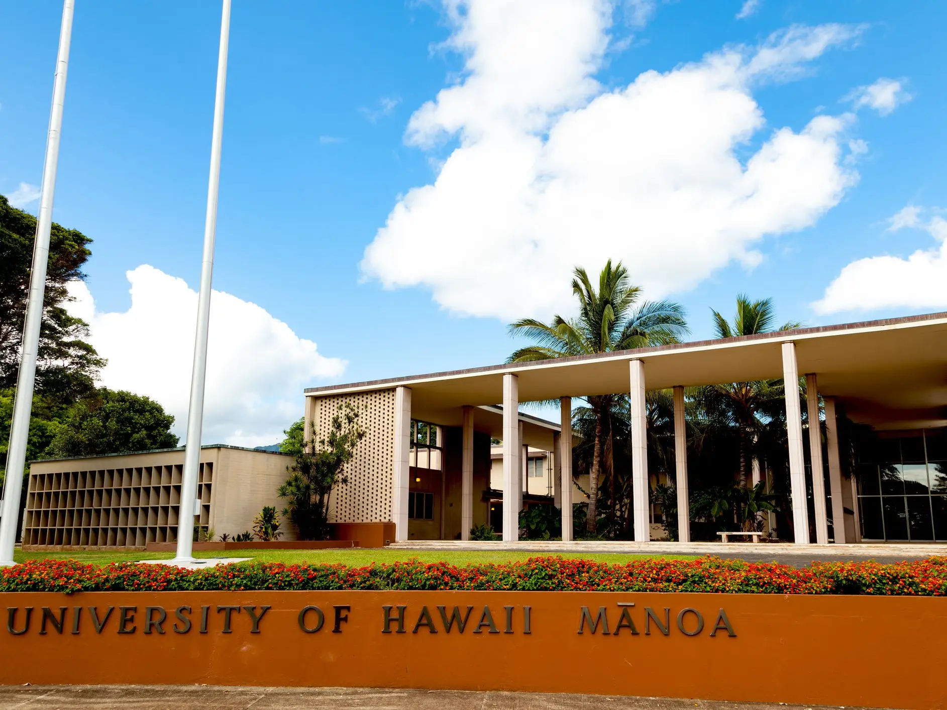 University of Hawaii Mānoa campus sign stands before a modern building with palm trees and a bright blue sky.