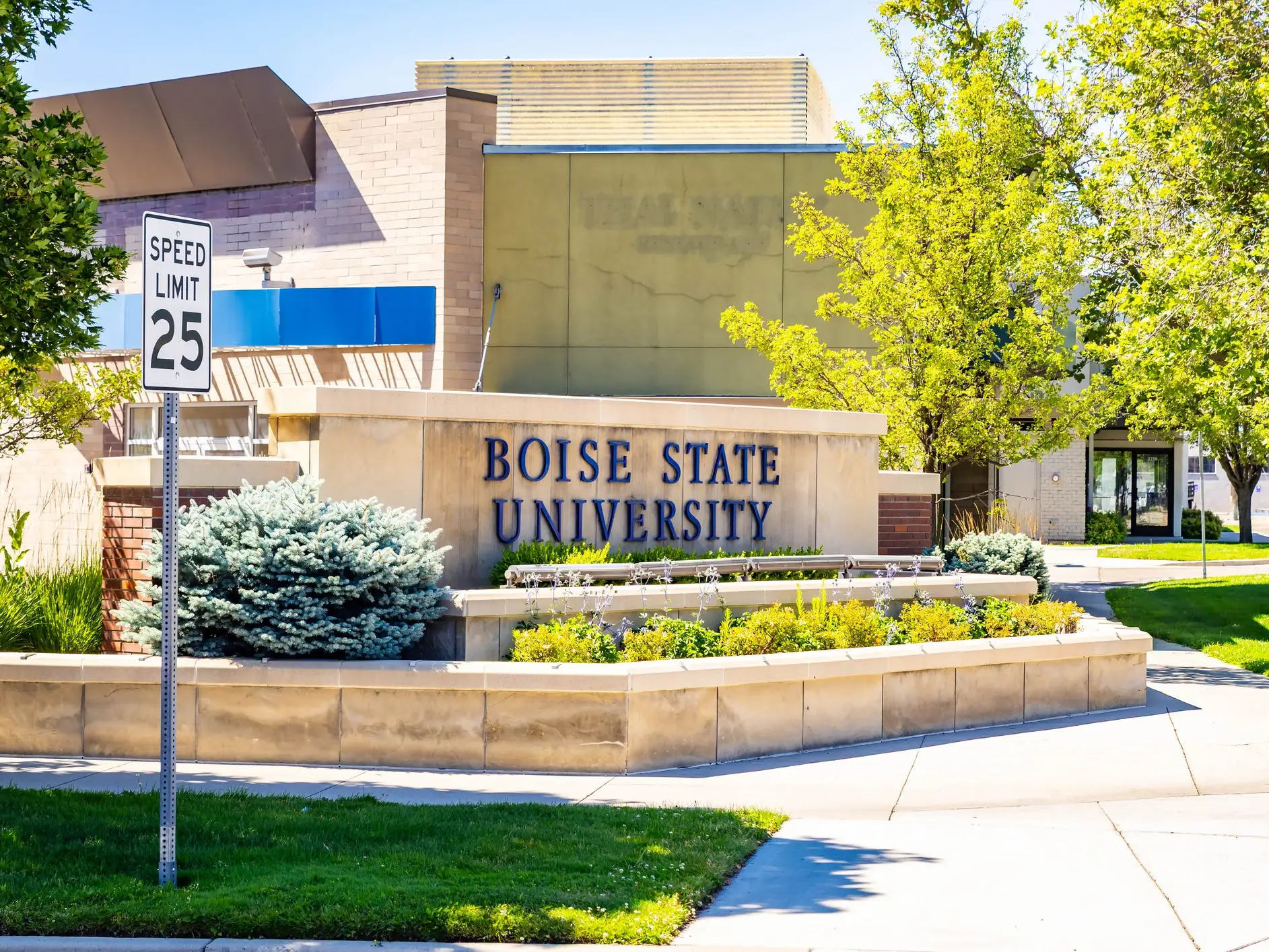 Boise State University entrance sign stands beside a sidewalk, landscaping, trees, and a SPEED LIMIT 25 road sign.