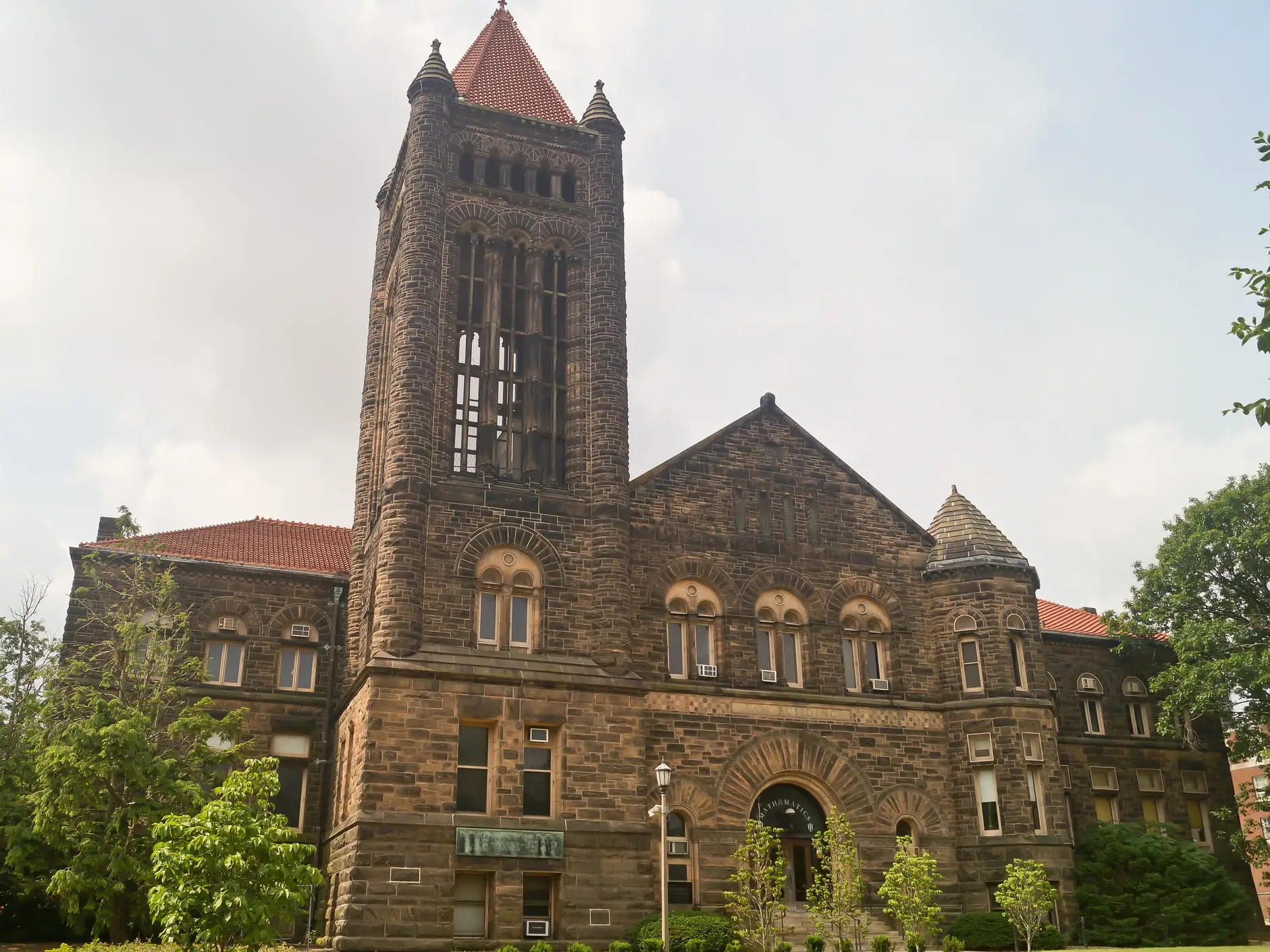 A University of Illinois Urbana-Champaign building rises behind trees, with a stone tower and a red-tile roof.