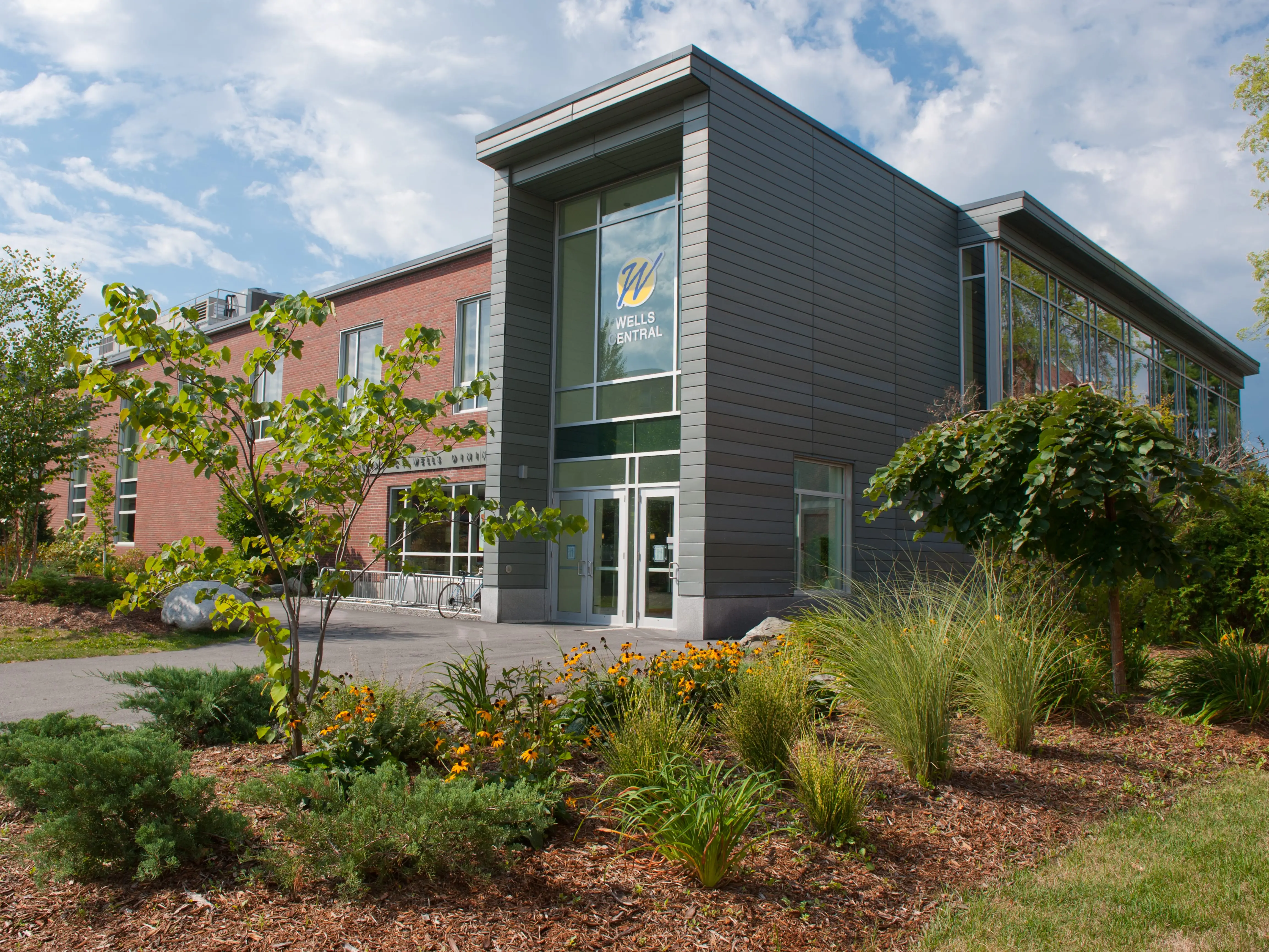 University of Maine building exterior with glass entrance, landscaped gardens, and trees under a partly cloudy sky.