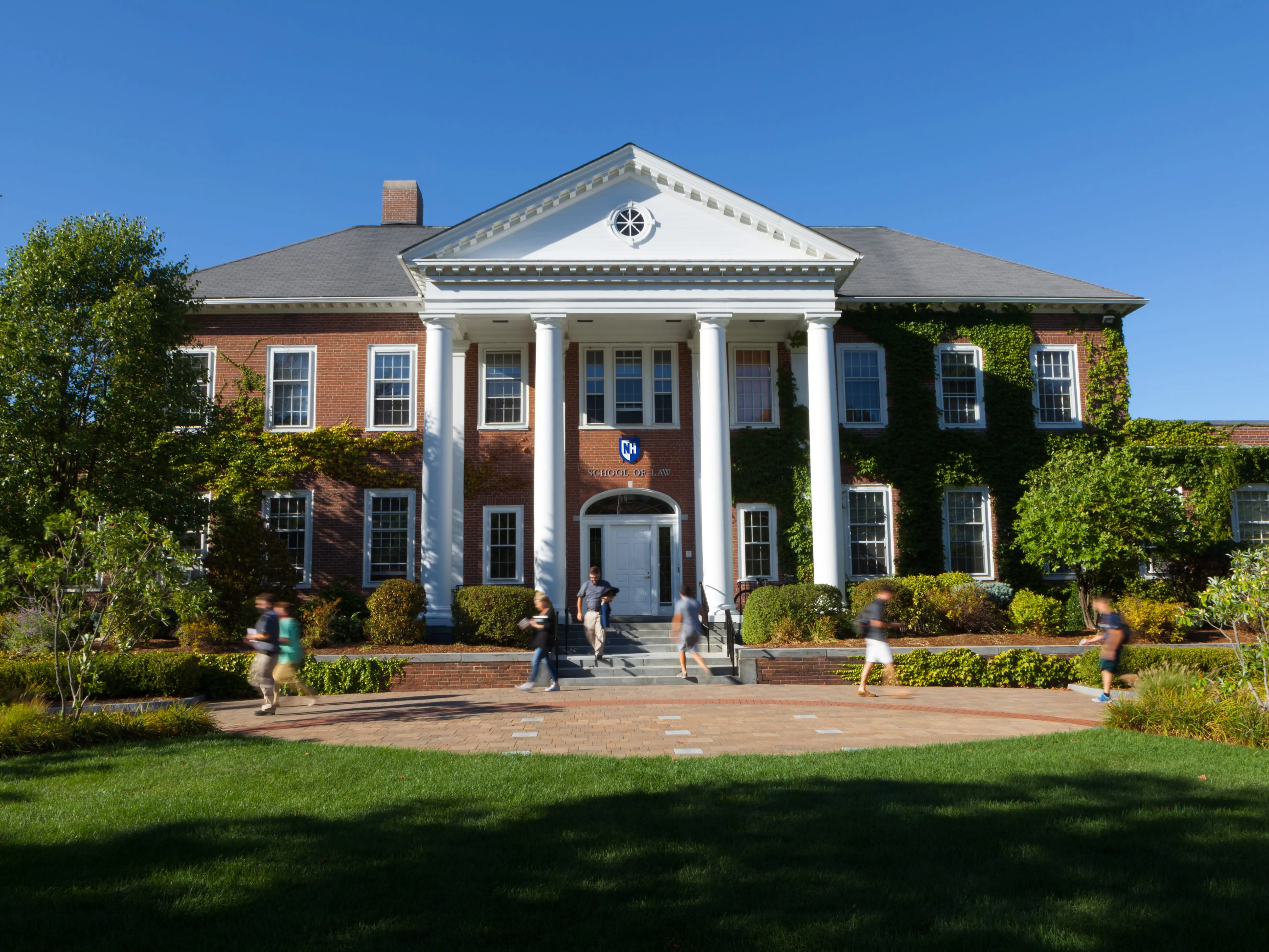 Red brick campus building with white columns, ivy, and people walking across a sunny lawn.