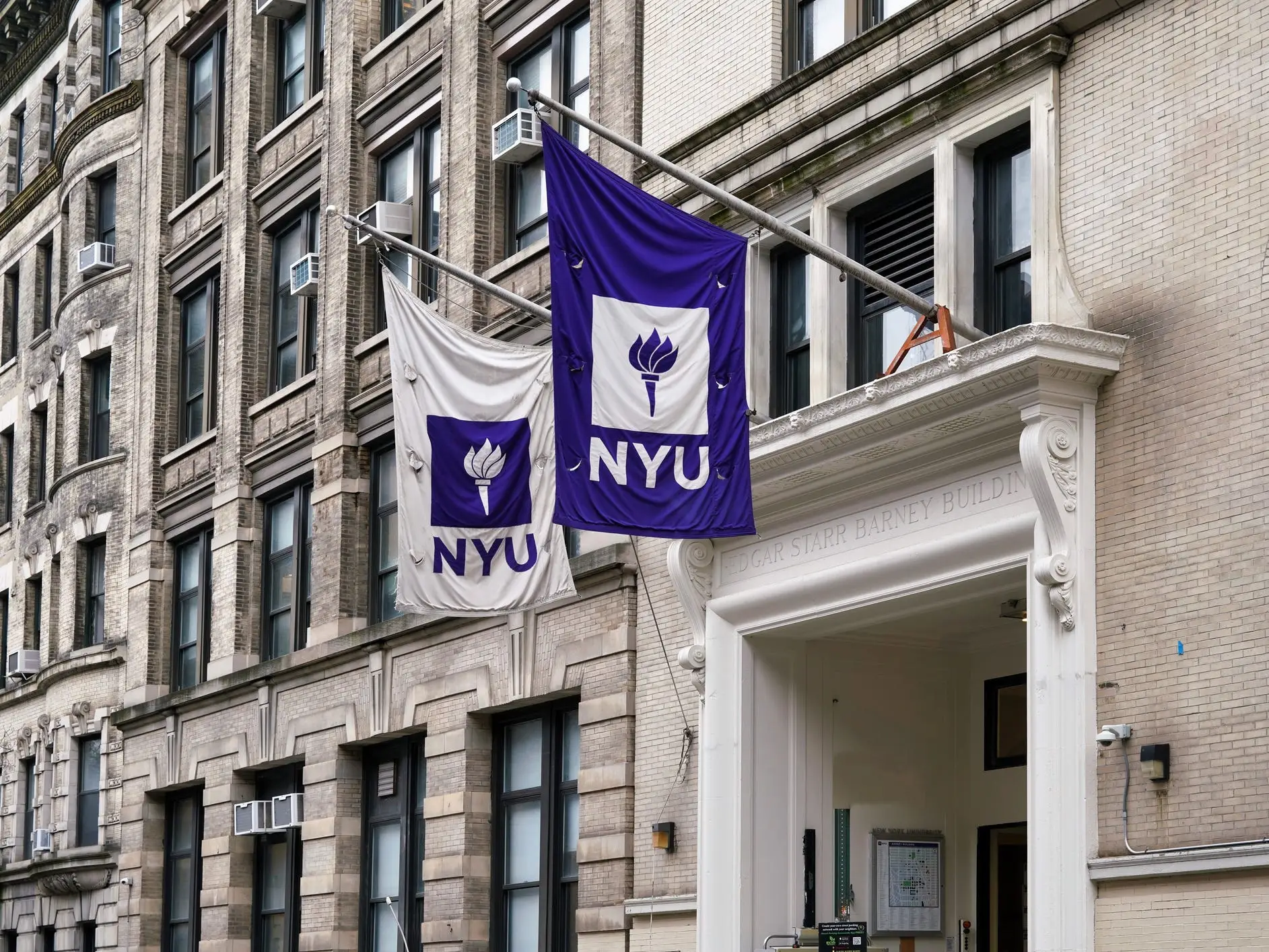 NYU flags hang above an ornate entrance on a multi-story brick campus building.