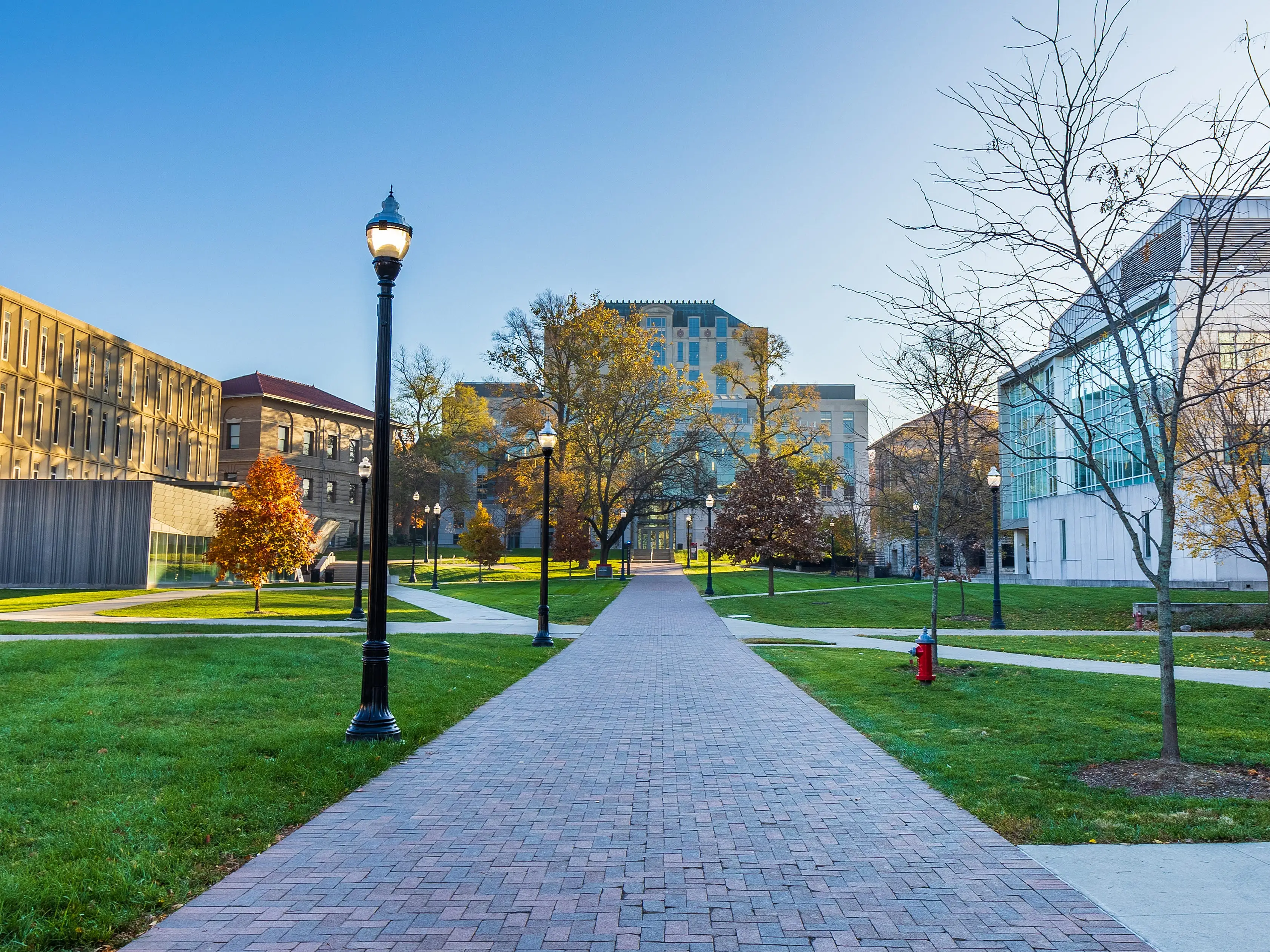 Brick walkway lined with lamp posts runs through a landscaped campus courtyard between academic buildings.