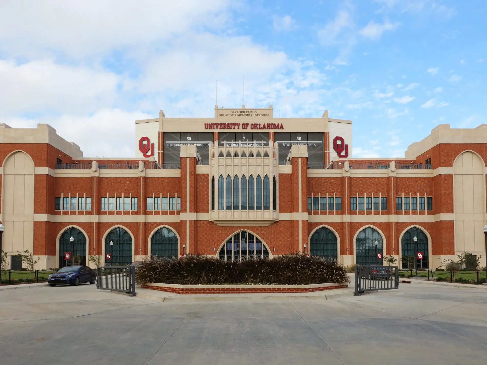 University of Oklahoma stadium façade with OU logos, arched windows, gates, and parked cars under a cloudy sky.