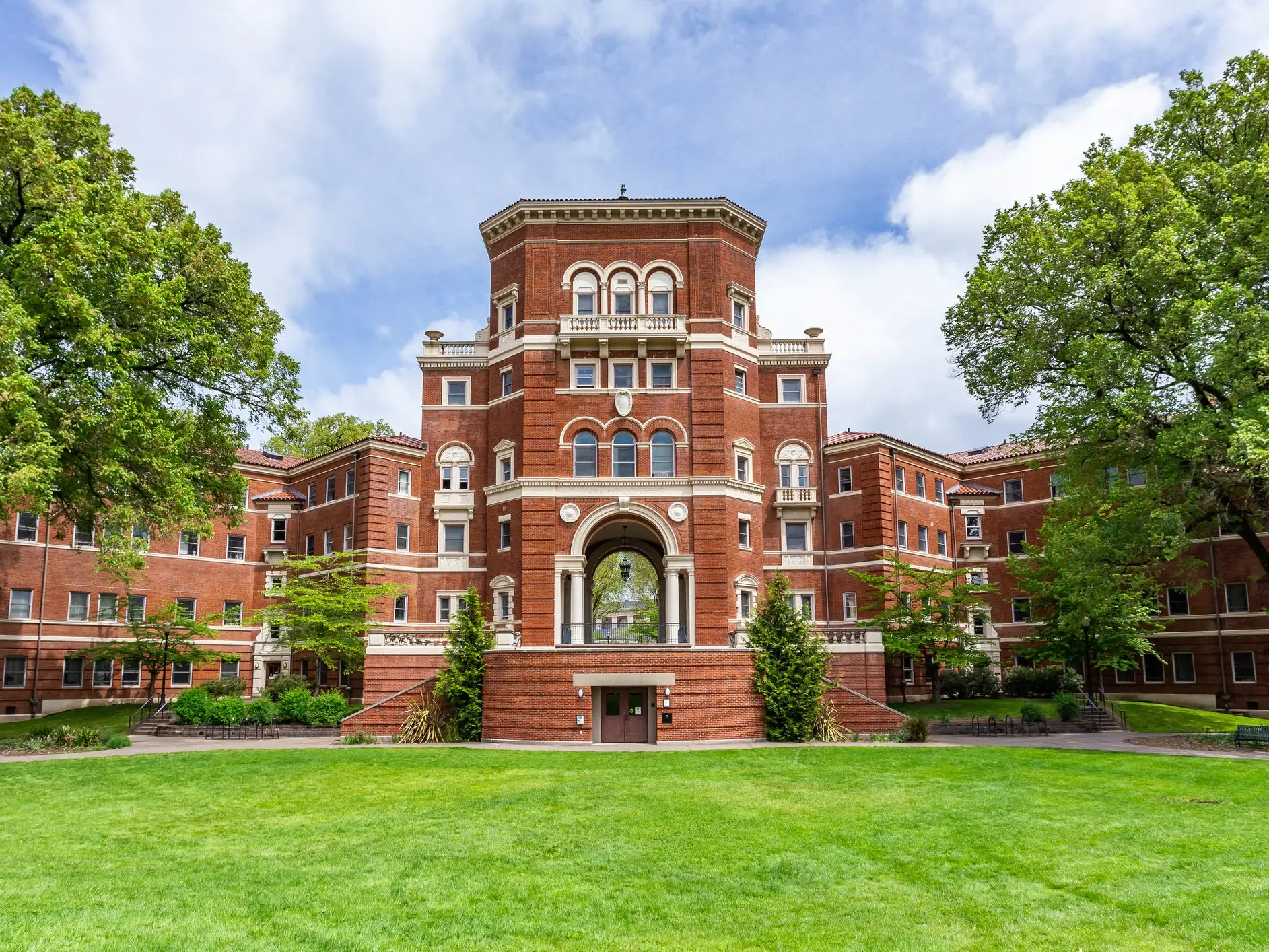 Red brick academic building with arched central entrance stands behind a green lawn and large trees.
