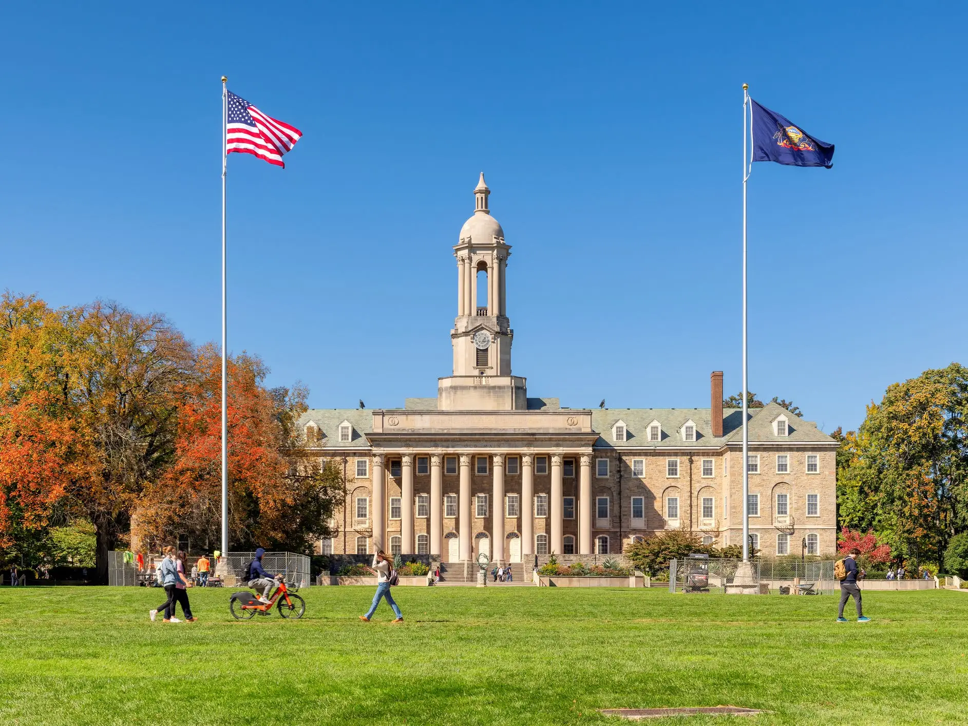 Old Main at Pennsylvania State University stands behind a lawn with flags and pedestrians on a clear autumn day.