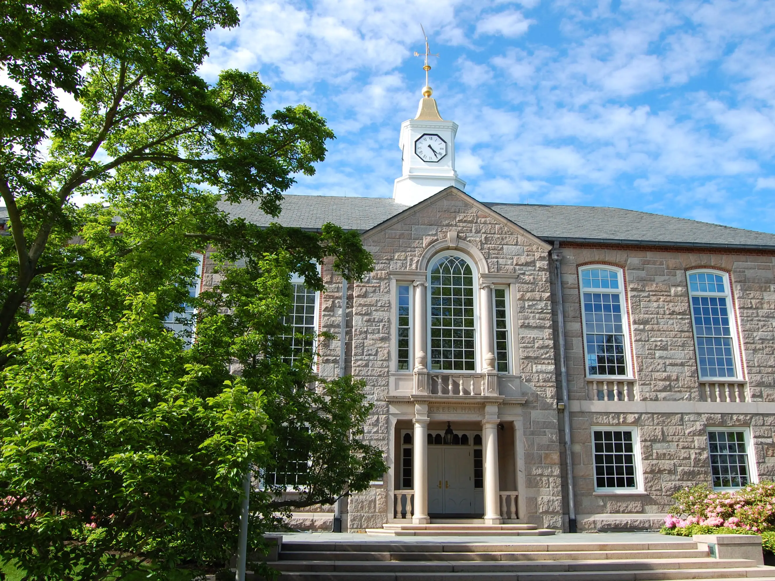 Stone academic-style building with a clock cupola stands behind trees under a partly cloudy blue sky.