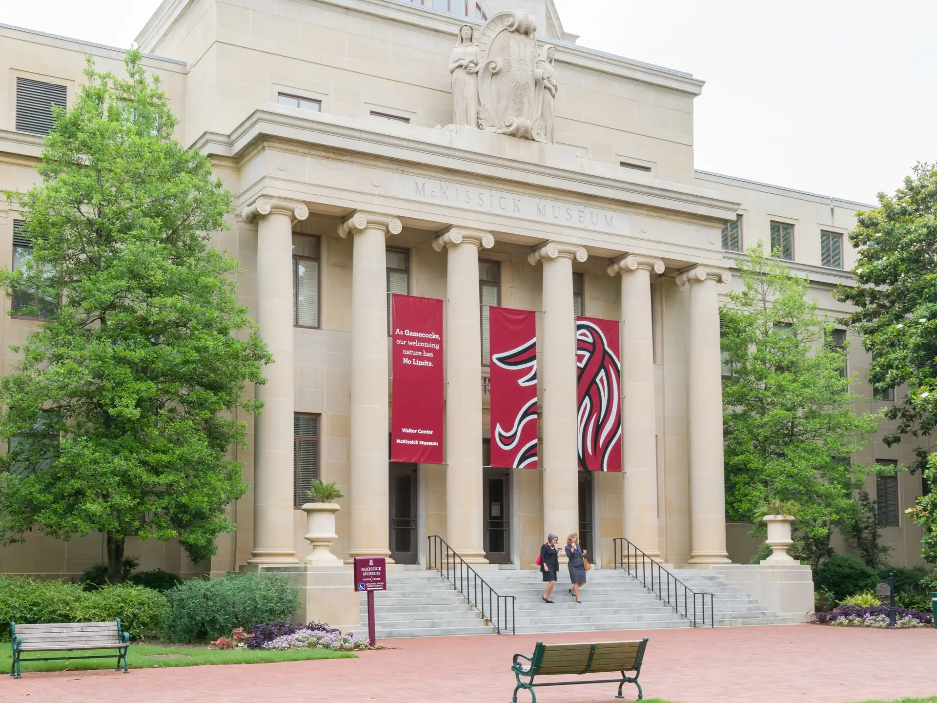 McKissick Museum entrance with tall columns, red banners, trees, benches, and two people on the front steps.