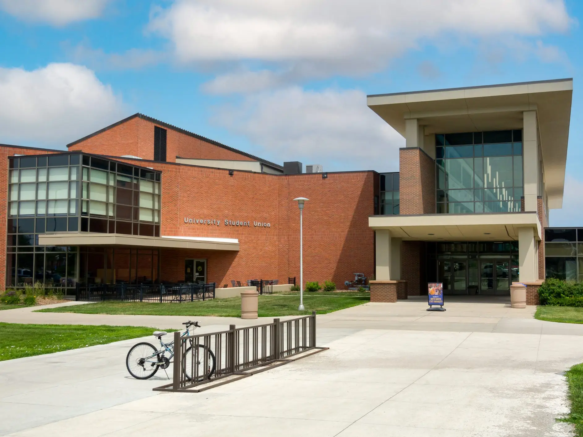 Student Union building with glass entrance, walkway, lawn, bike rack, and bicycle under a partly cloudy sky.