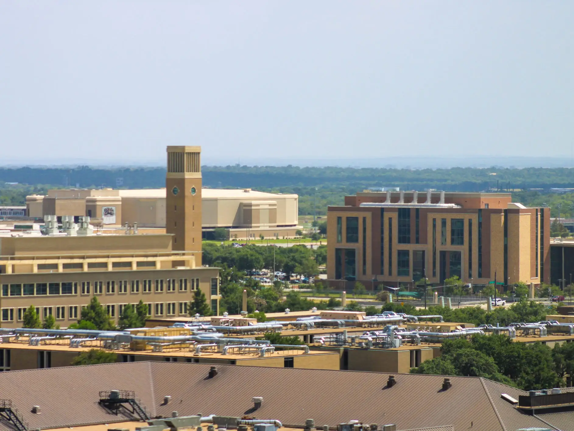 Large campus buildings and rooftops with a tower are seen from an elevated viewpoint under a hazy sky.