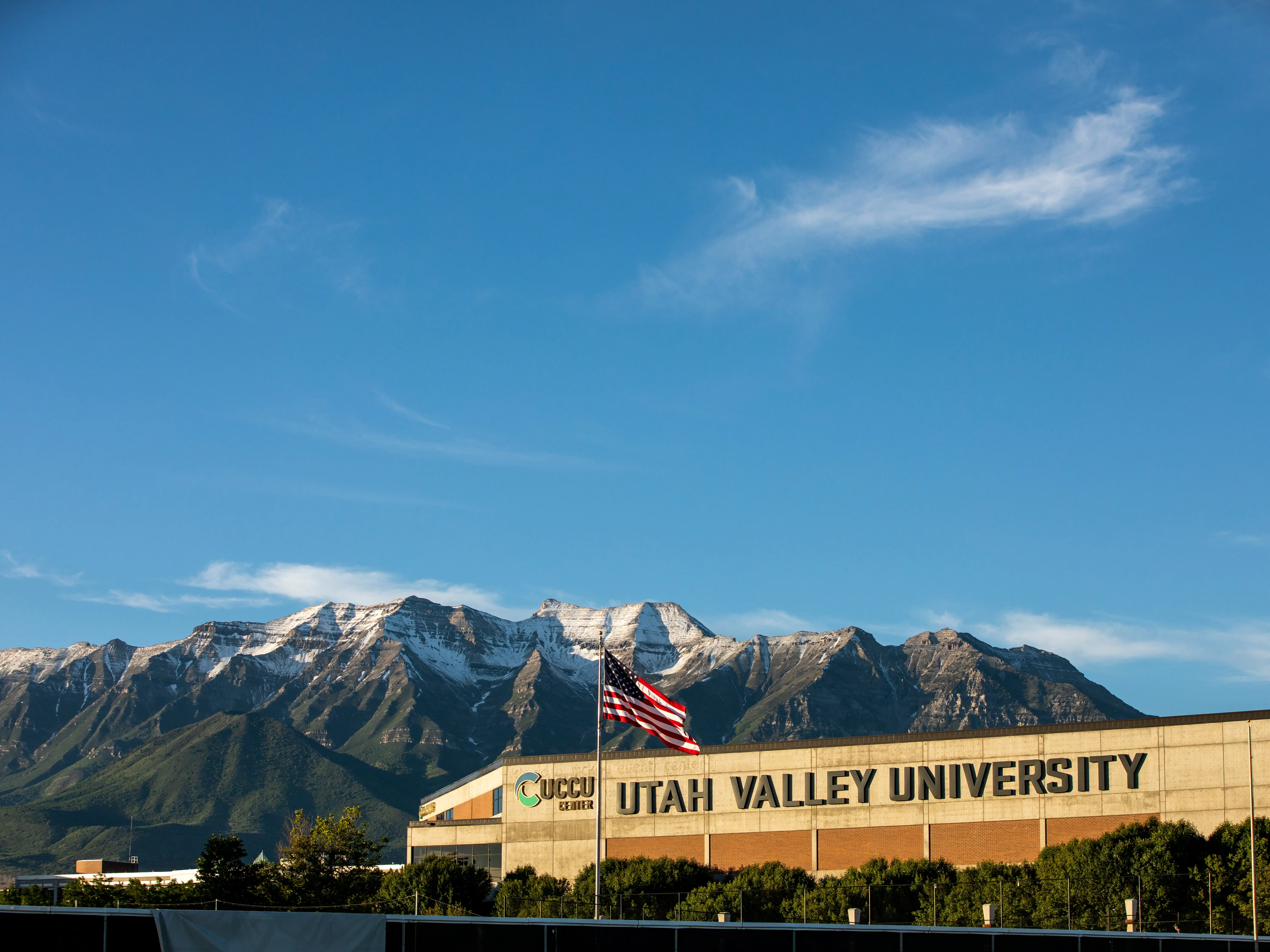 Utah Valley University building with an American flag stands before snow-dusted mountains under a blue sky.
