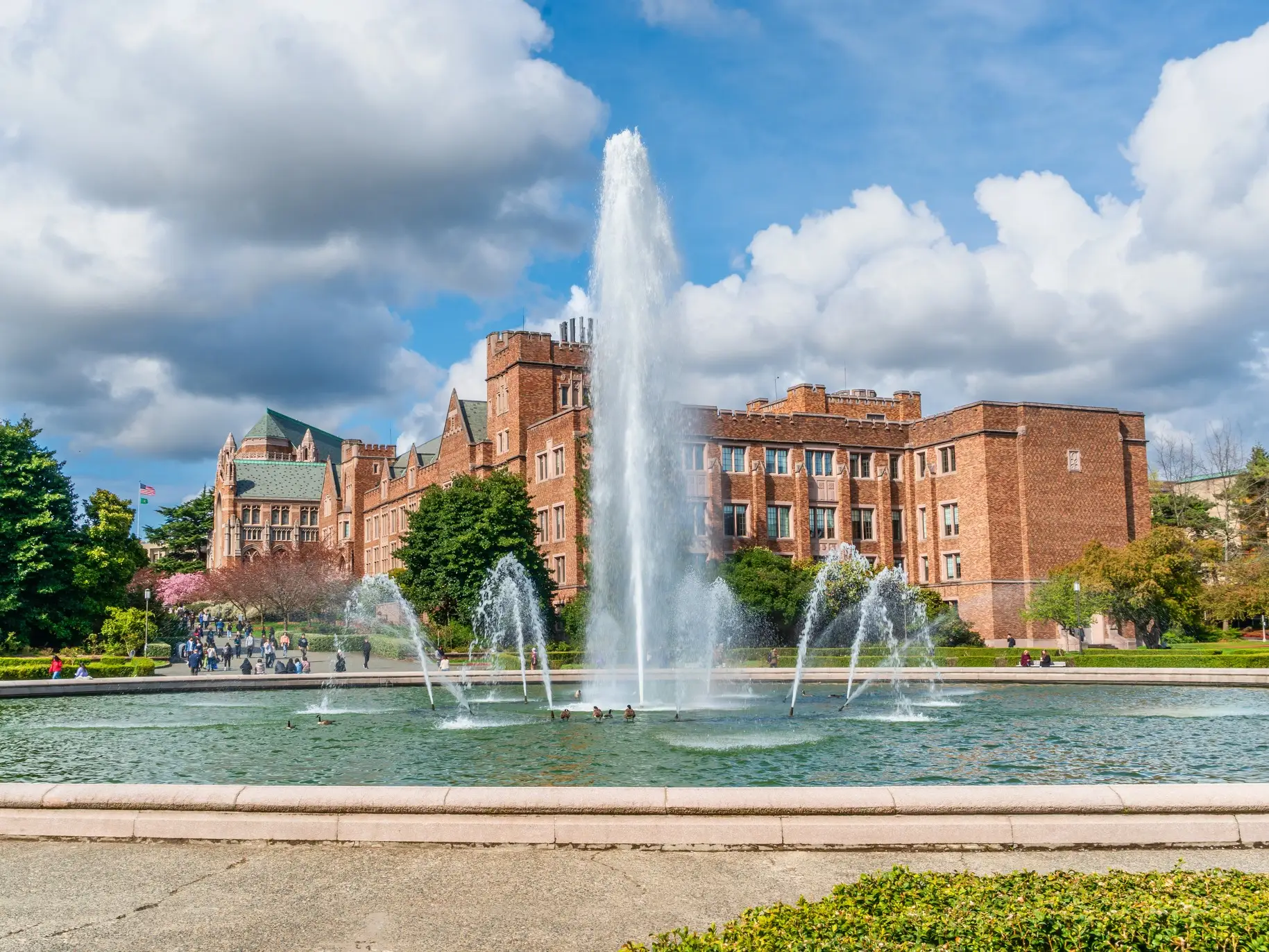 A large campus fountain sprays in front of red brick buildings under a partly cloudy sky.