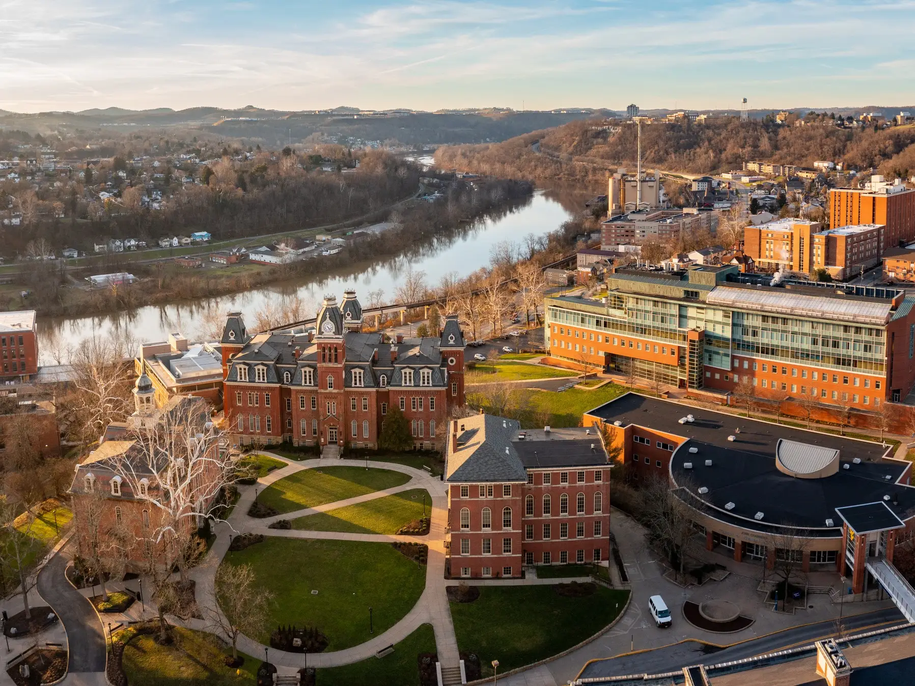 Aerial view of a university campus with brick buildings beside a river and wooded hills at sunset.