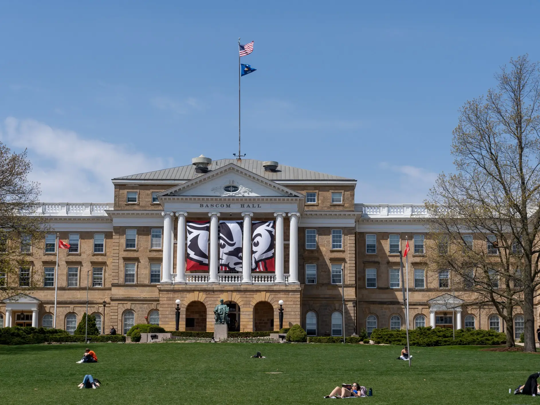 Bascom Hall with flags and a large badger banner overlooks people relaxing on a green campus lawn.
