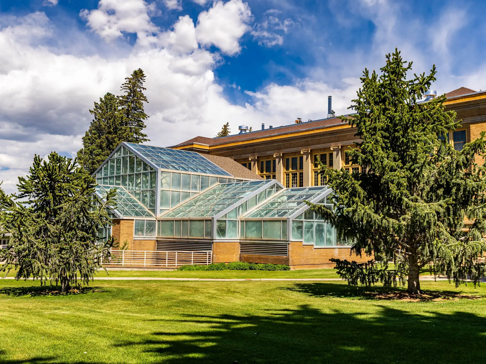 Glass greenhouse structure stands beside a brick building on a landscaped lawn under a partly cloudy sky.