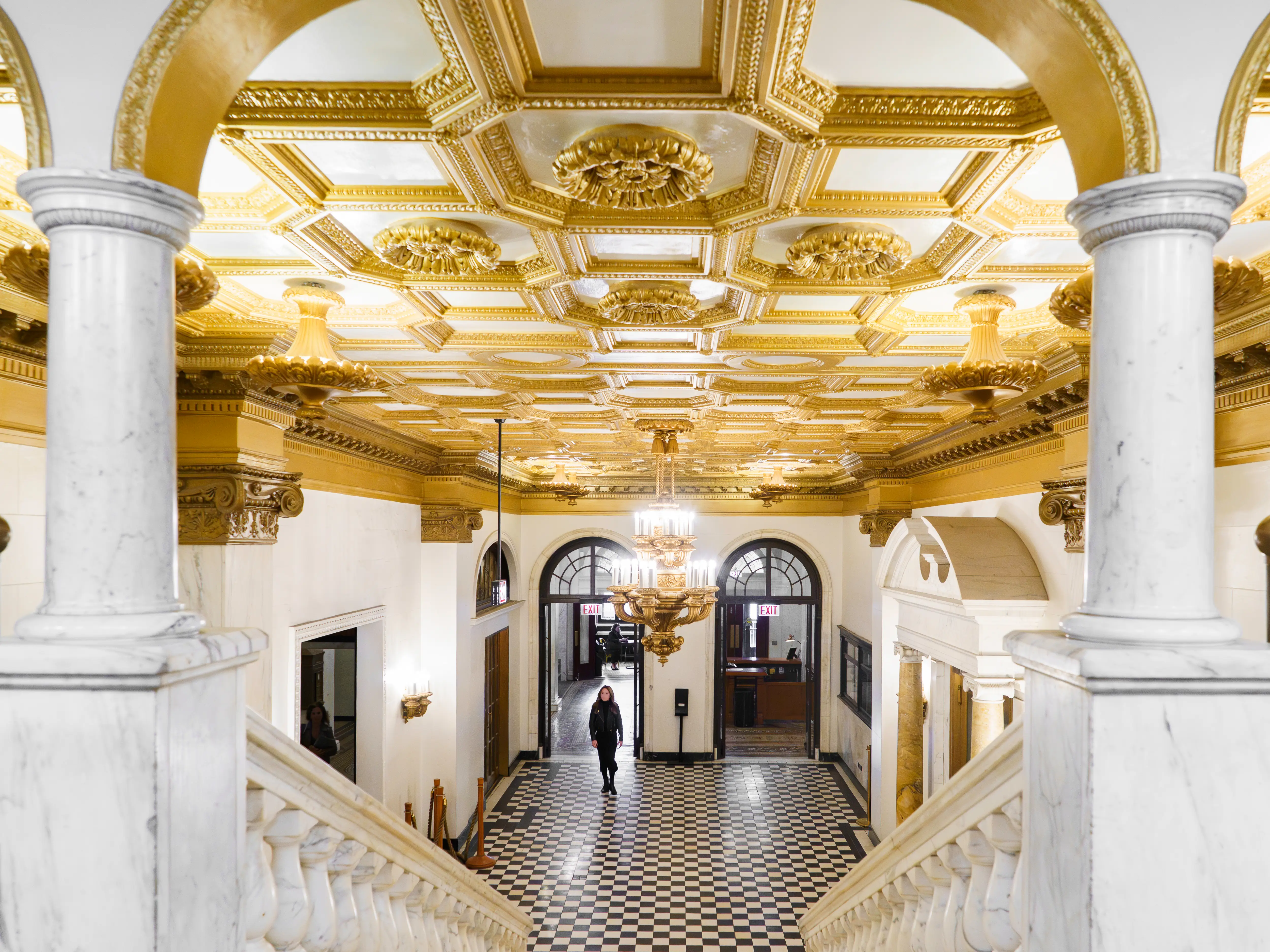 Ornate lobby with gold coffered ceiling, marble columns, chandeliers, staircases, and black-and-white checkered floor.