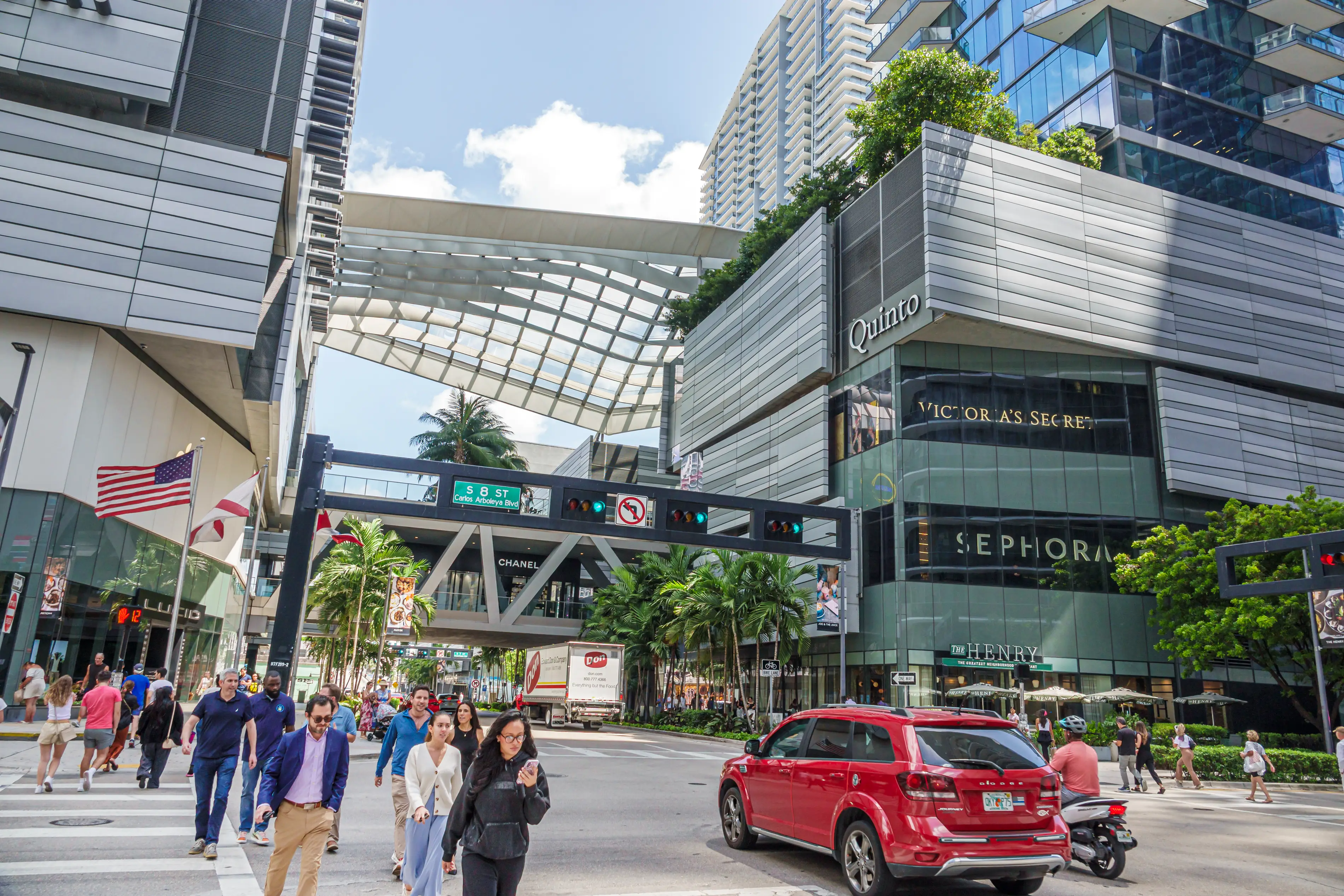 People walk through a busy downtown with palm trees in the background.