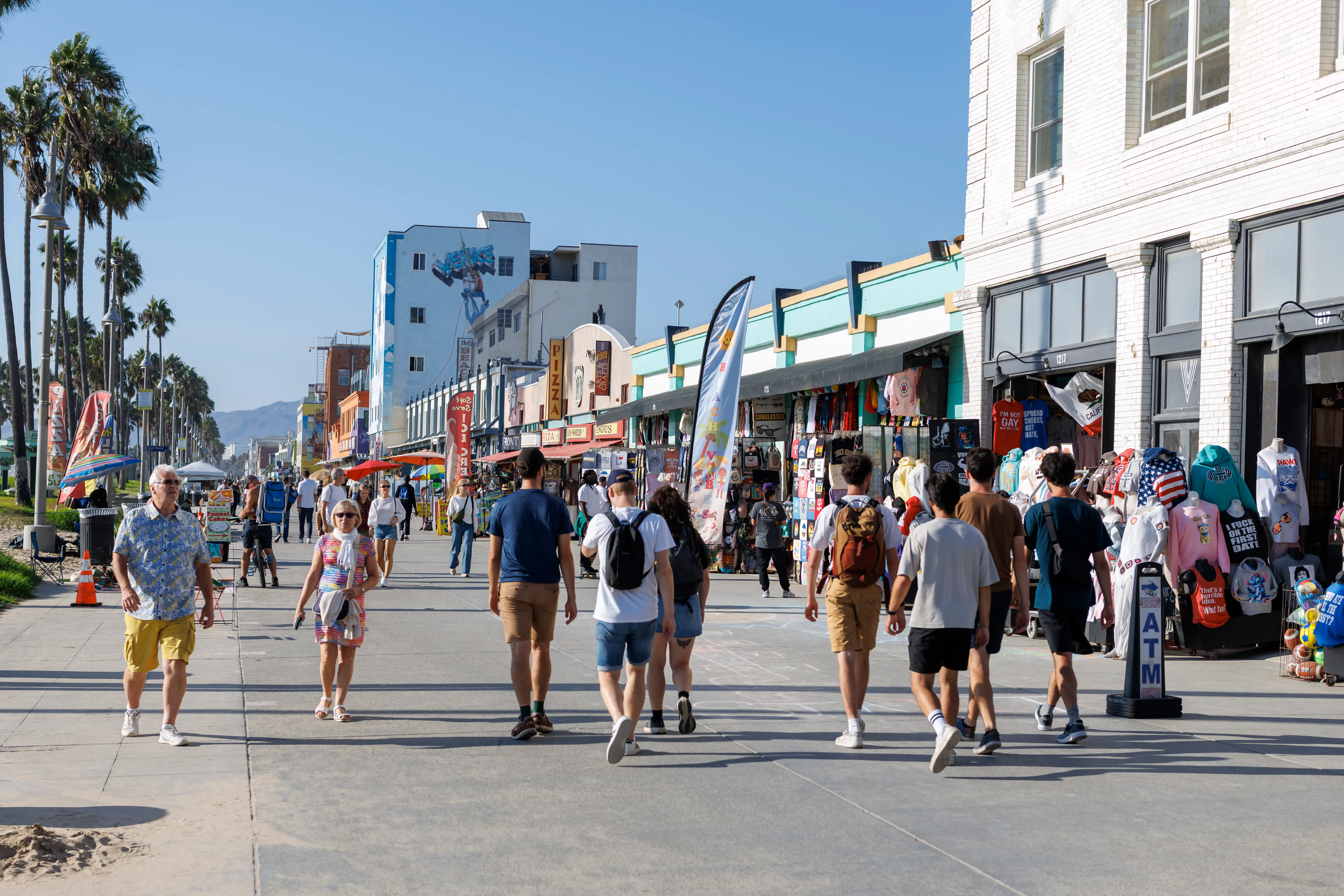 People walk along a sunny boardwalk with palm trees to the left.
