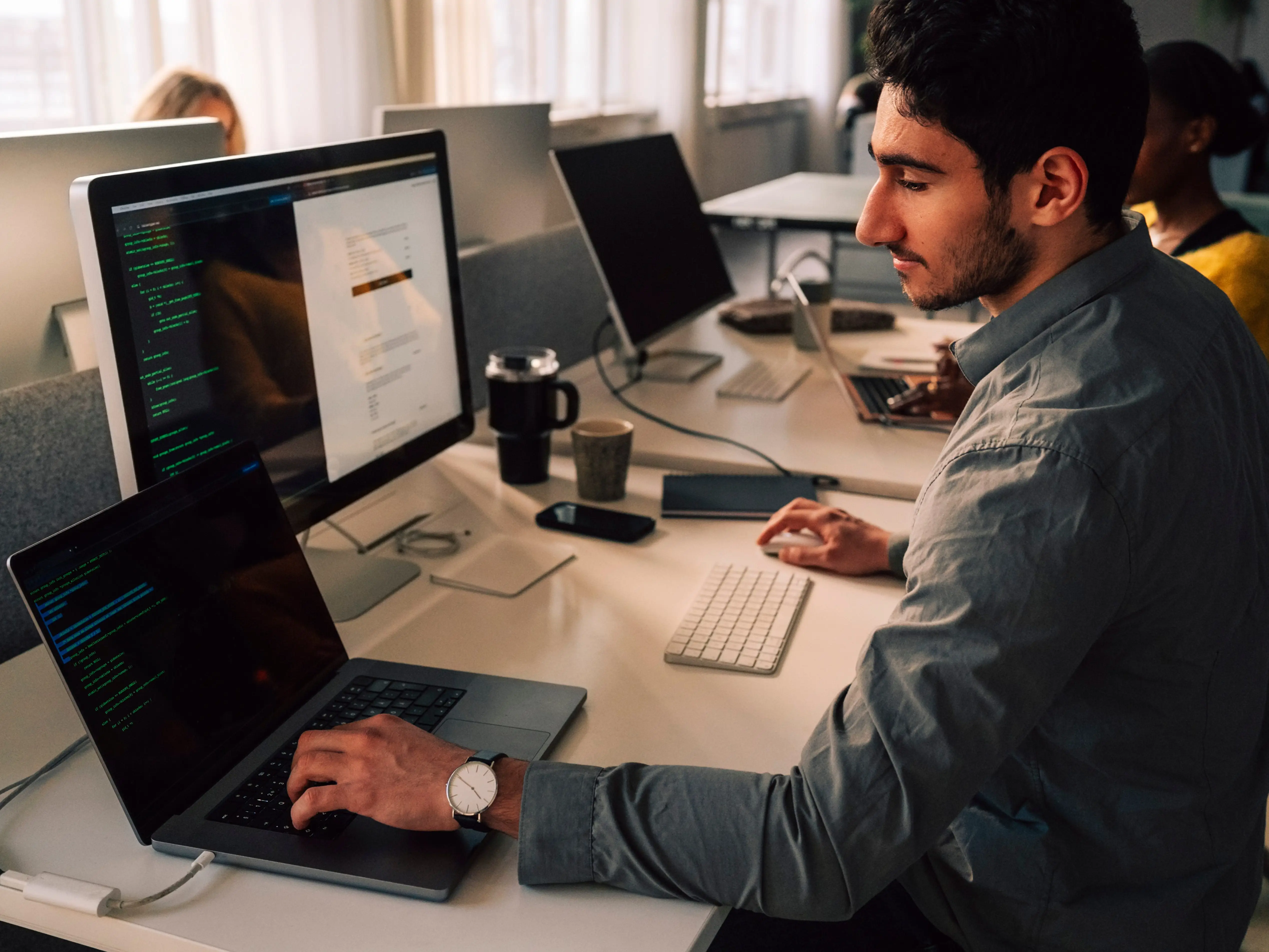 A person in an office works at a laptop and a desktop monitor with code displayed