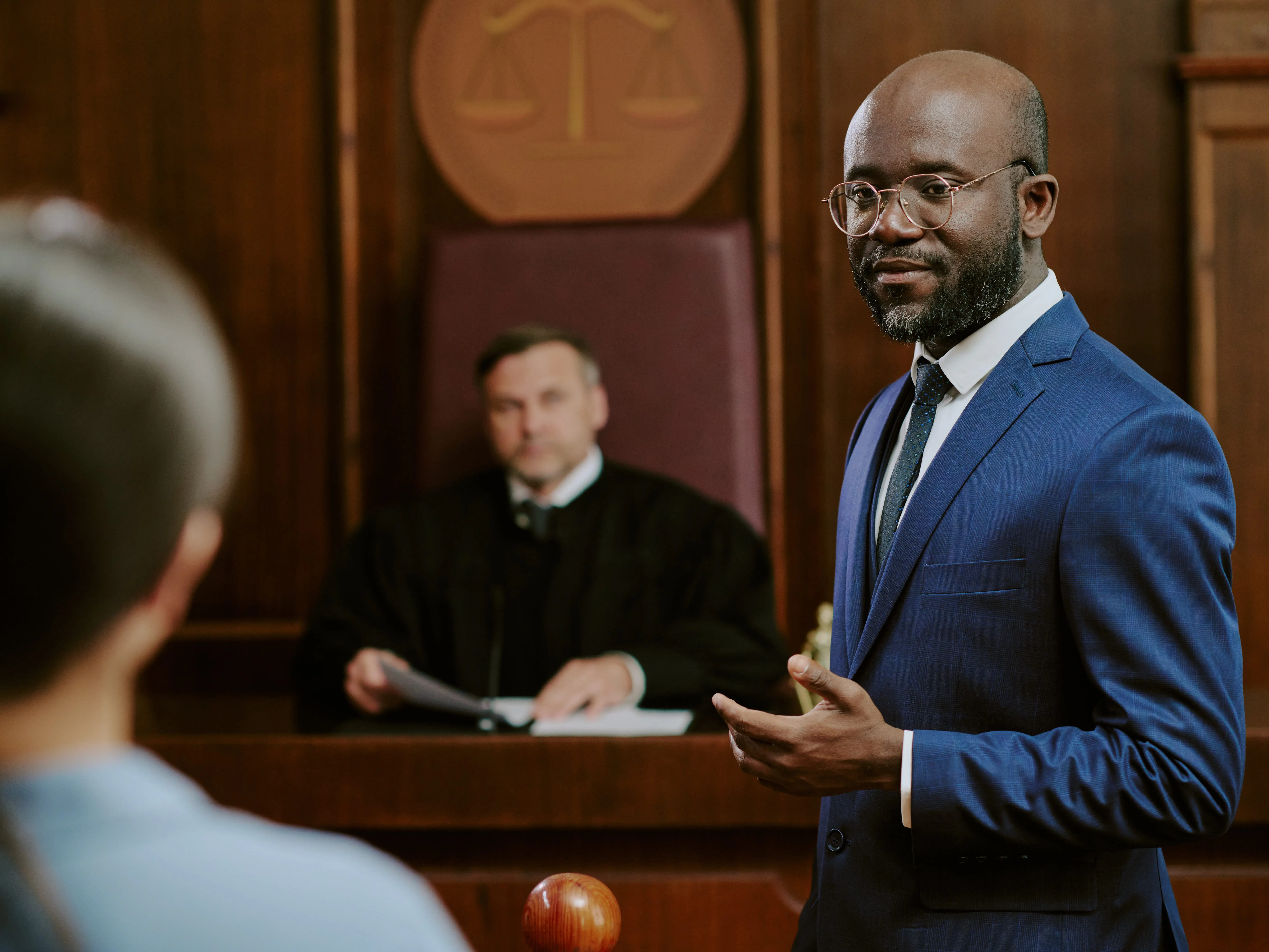 A person in a blue suit is looking at someone in a courtroom, and  a judge is seated