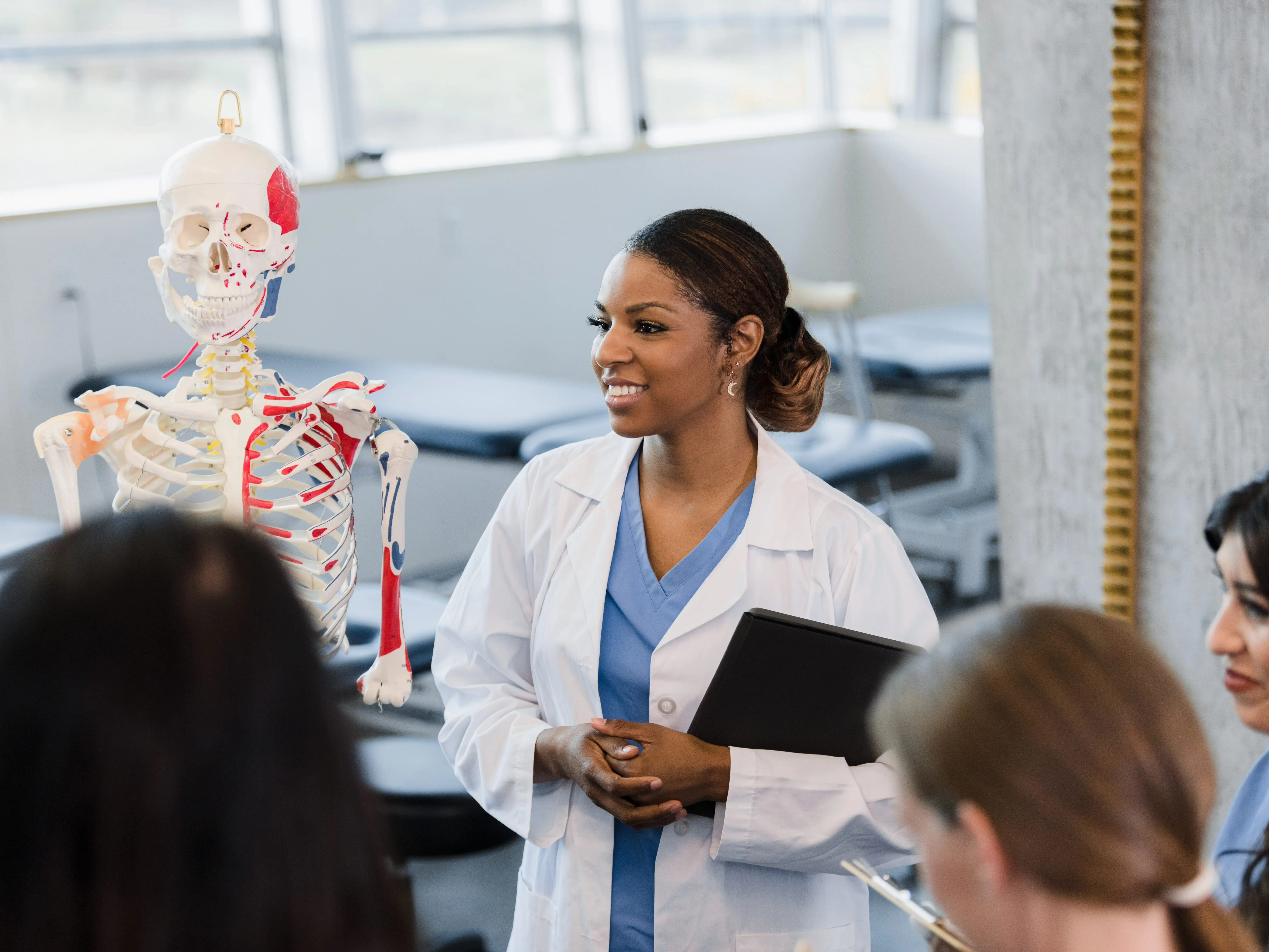 A medical trainer in a lab coat discusses a skeleton model with students