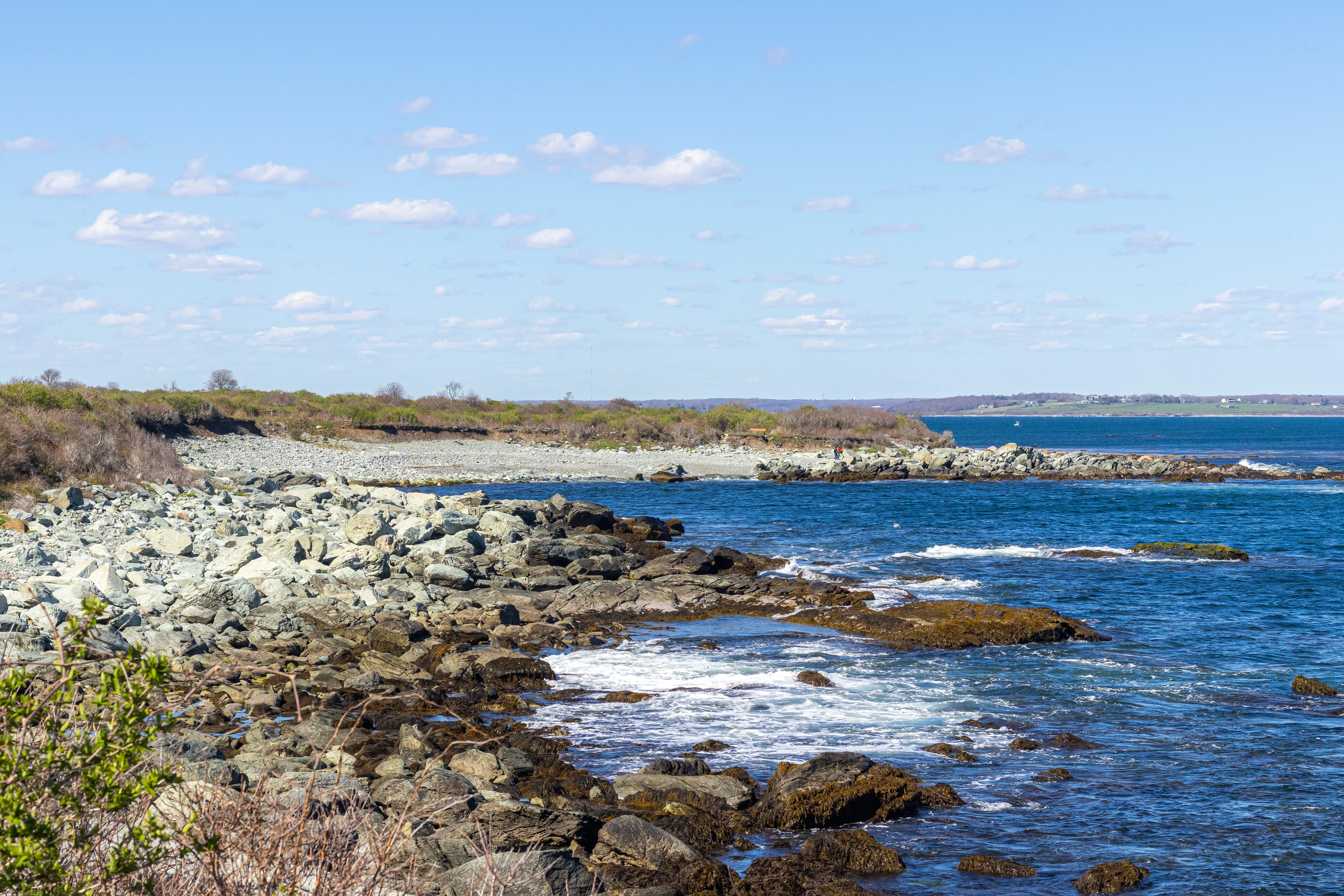 A rocky shoreline in Middletown, Rhode Island, curves along a blue ocean under a bright sky with scattered clouds.