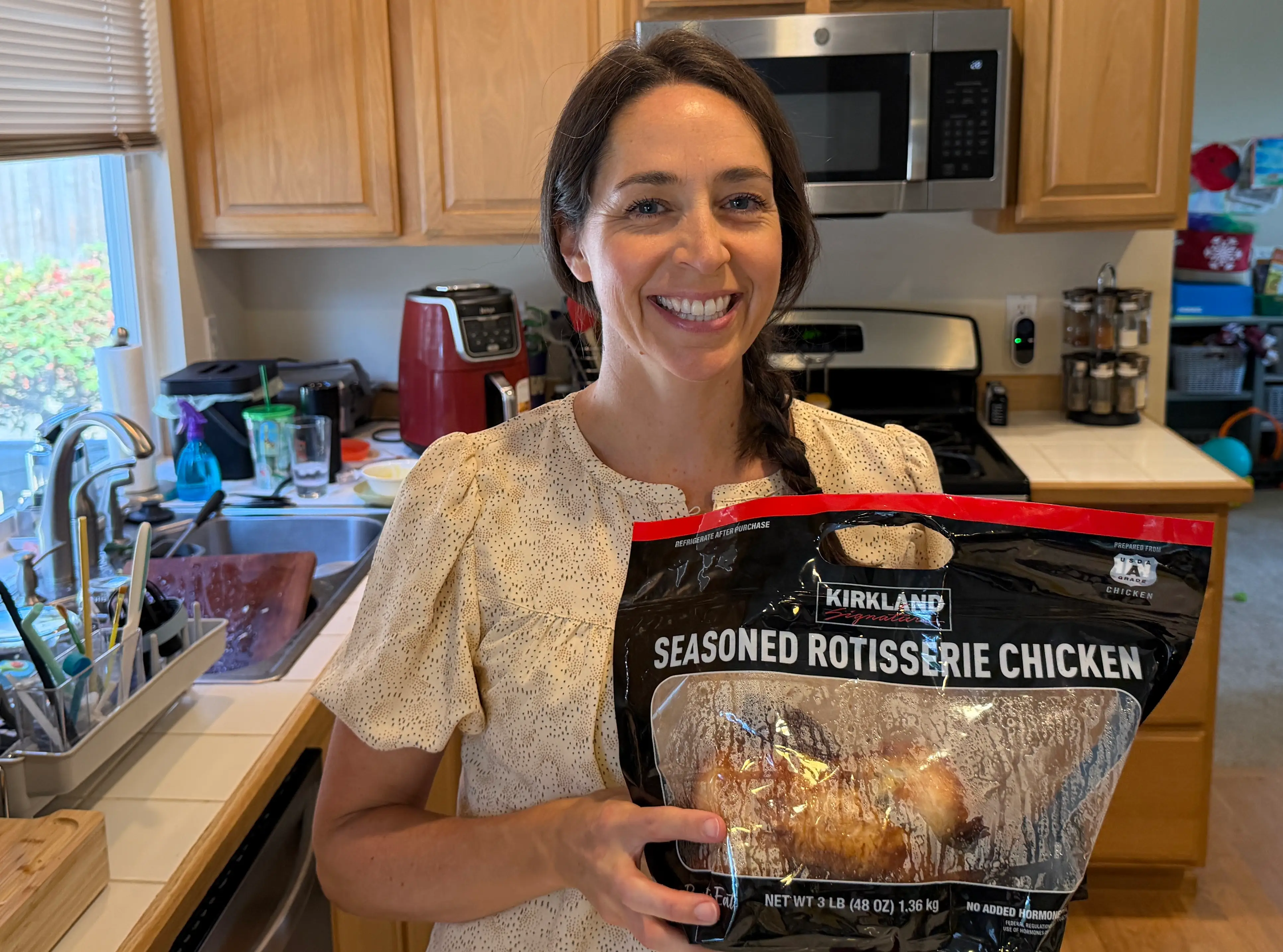 Woman smiling holding Kirkland Signature rotisserie chicken in kitchen
